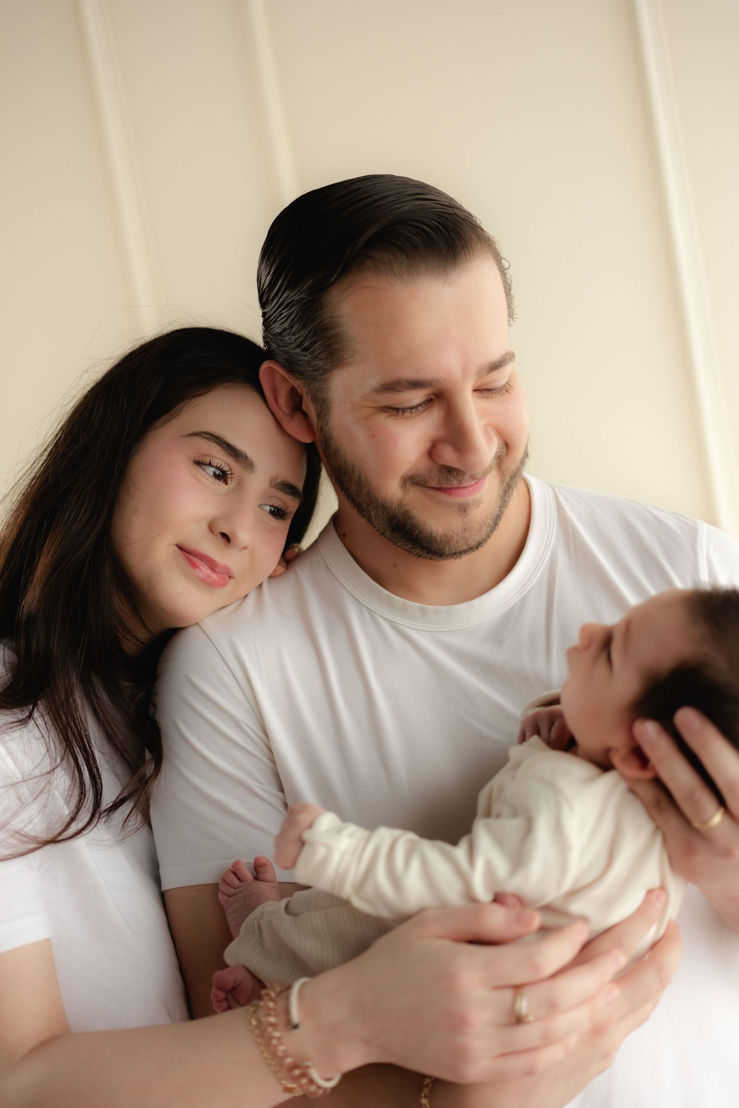 Happy young family with dark hair poses together, parents smiling while holding their newborn baby in white outfits.