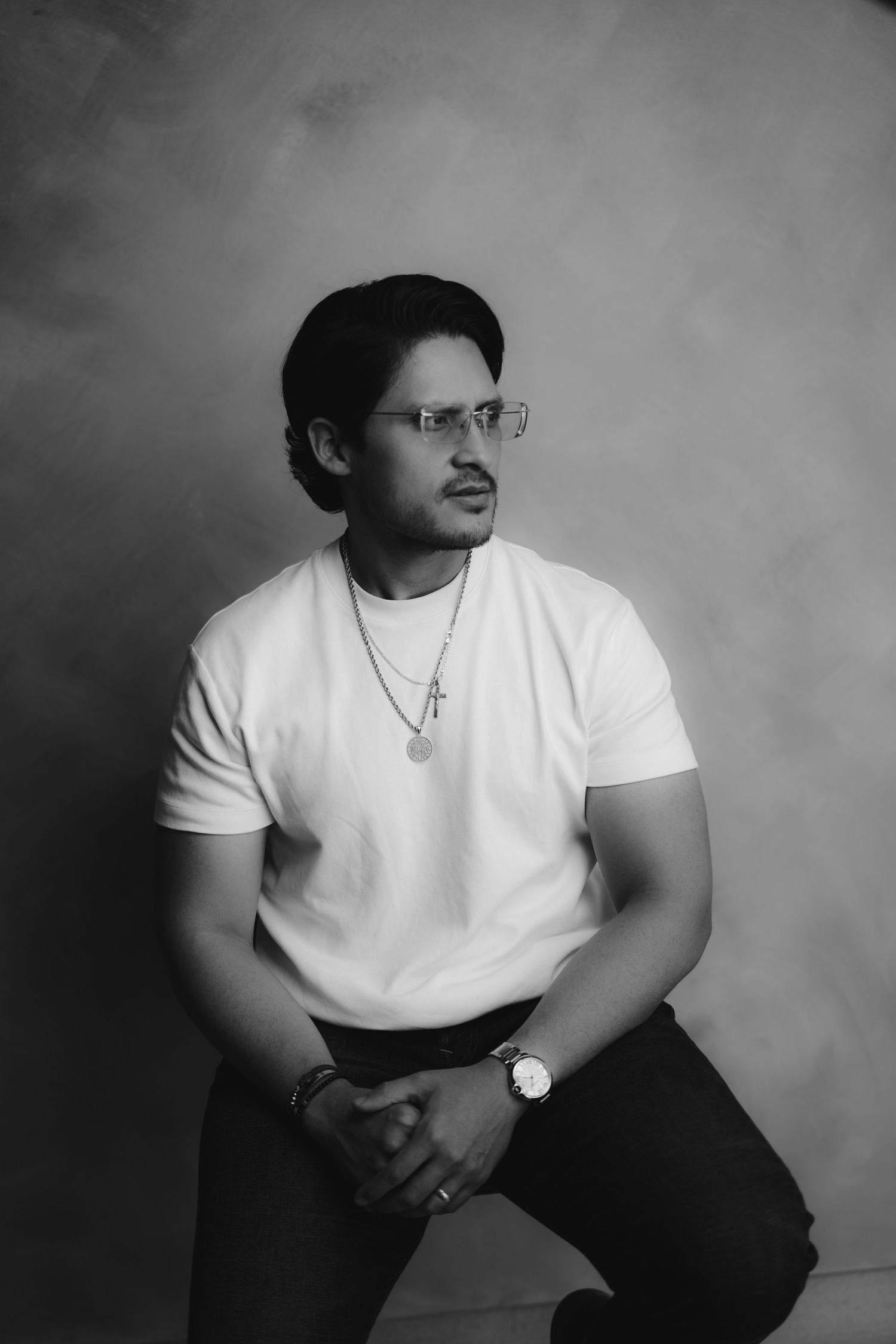 Stylish man in white t-shirt with cross necklace and glasses poses in black and white studio.