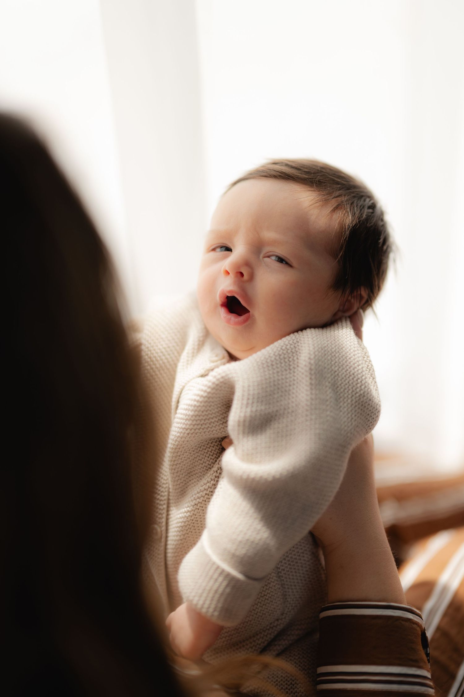 Adorable baby in a cream knit sweater yawning while being held, bathed in soft natural window light.