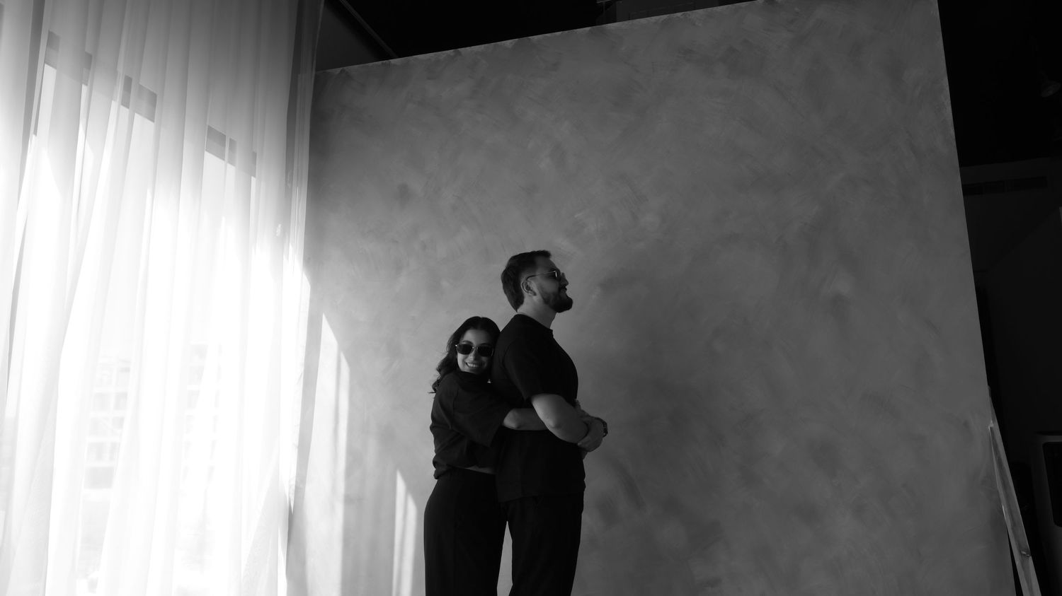 Black and white photo of a couple standing together near a window with sheer curtains.