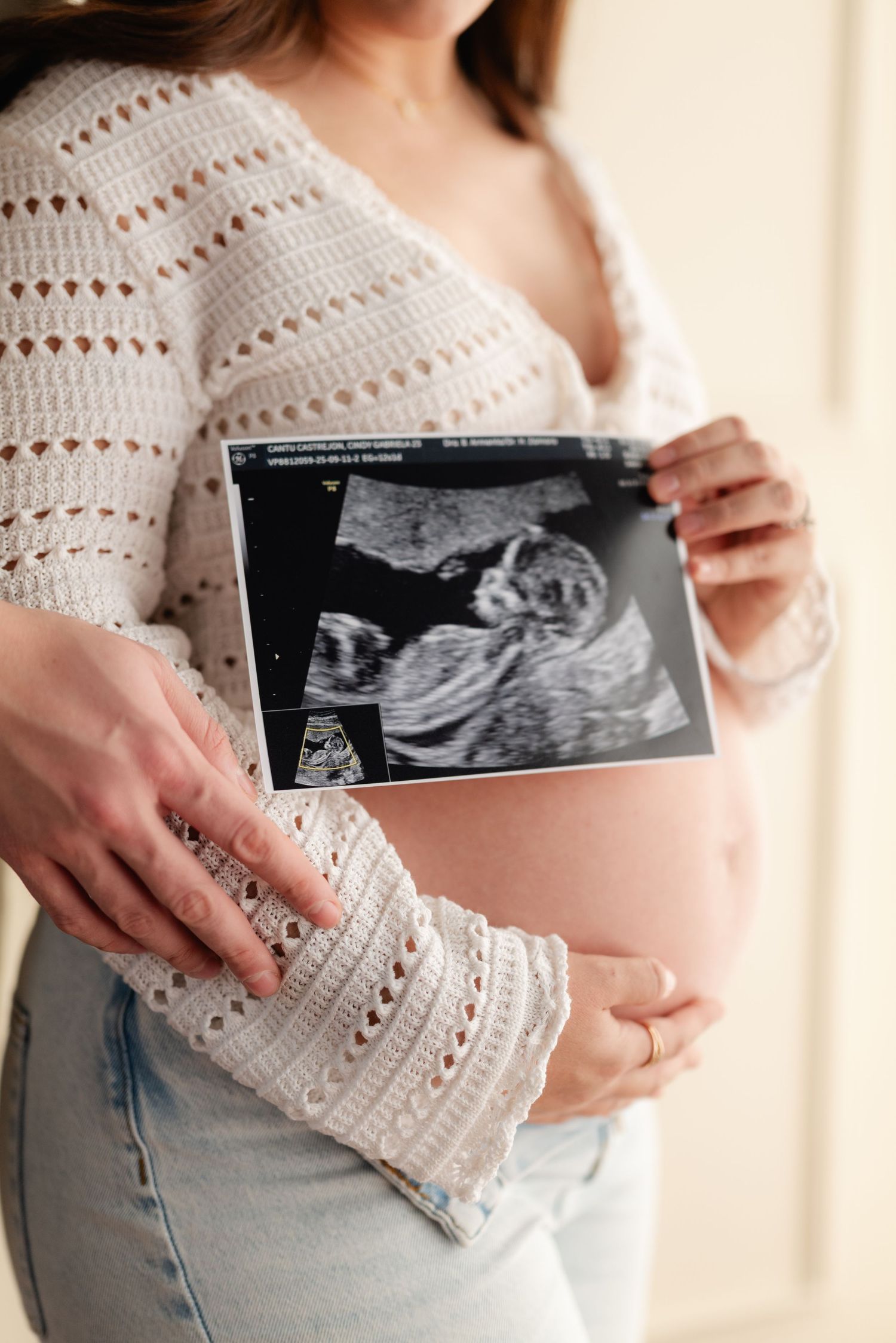 Pregnant woman in white crochet top and jeans holds ultrasound scan image next to her bare baby bump.