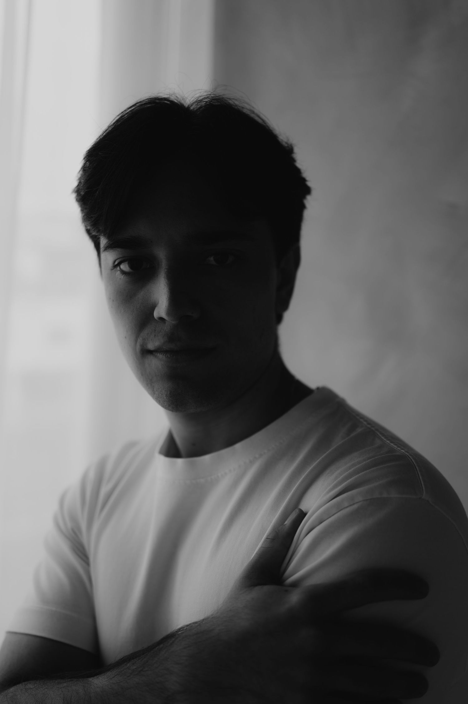 Brooding black and white portrait of a young man with crossed arms posing near a curtained window.