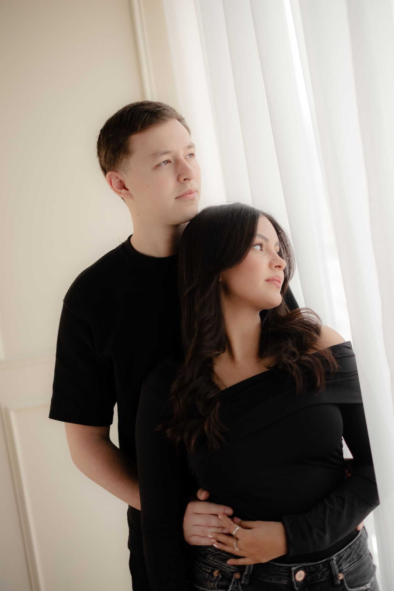 Elegant couple dressed in black embracing near sheer white curtains in a bright minimalist room.