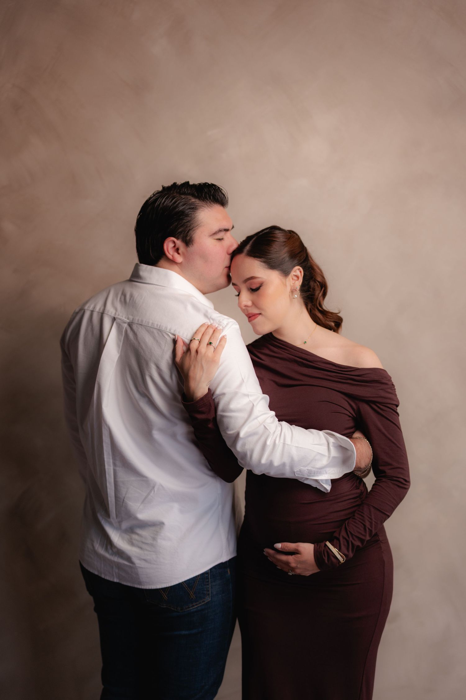 Man in white shirt kisses pregnant woman on forehead as she leans into him, wearing a burgundy maternity dress.