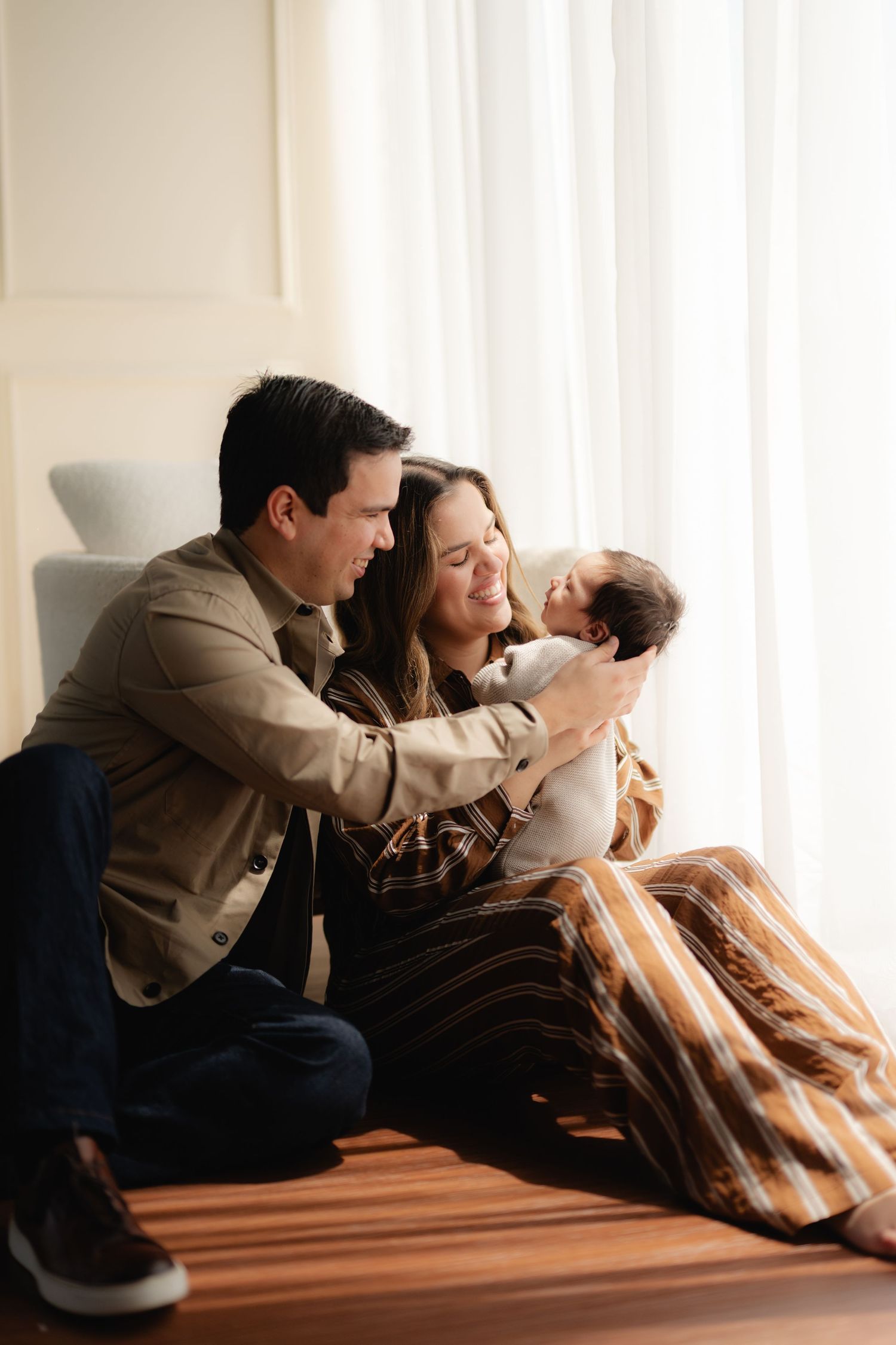 A couple sits on a hardwood floor by a window, lovingly gazing at their newborn baby wrapped in a blanket.