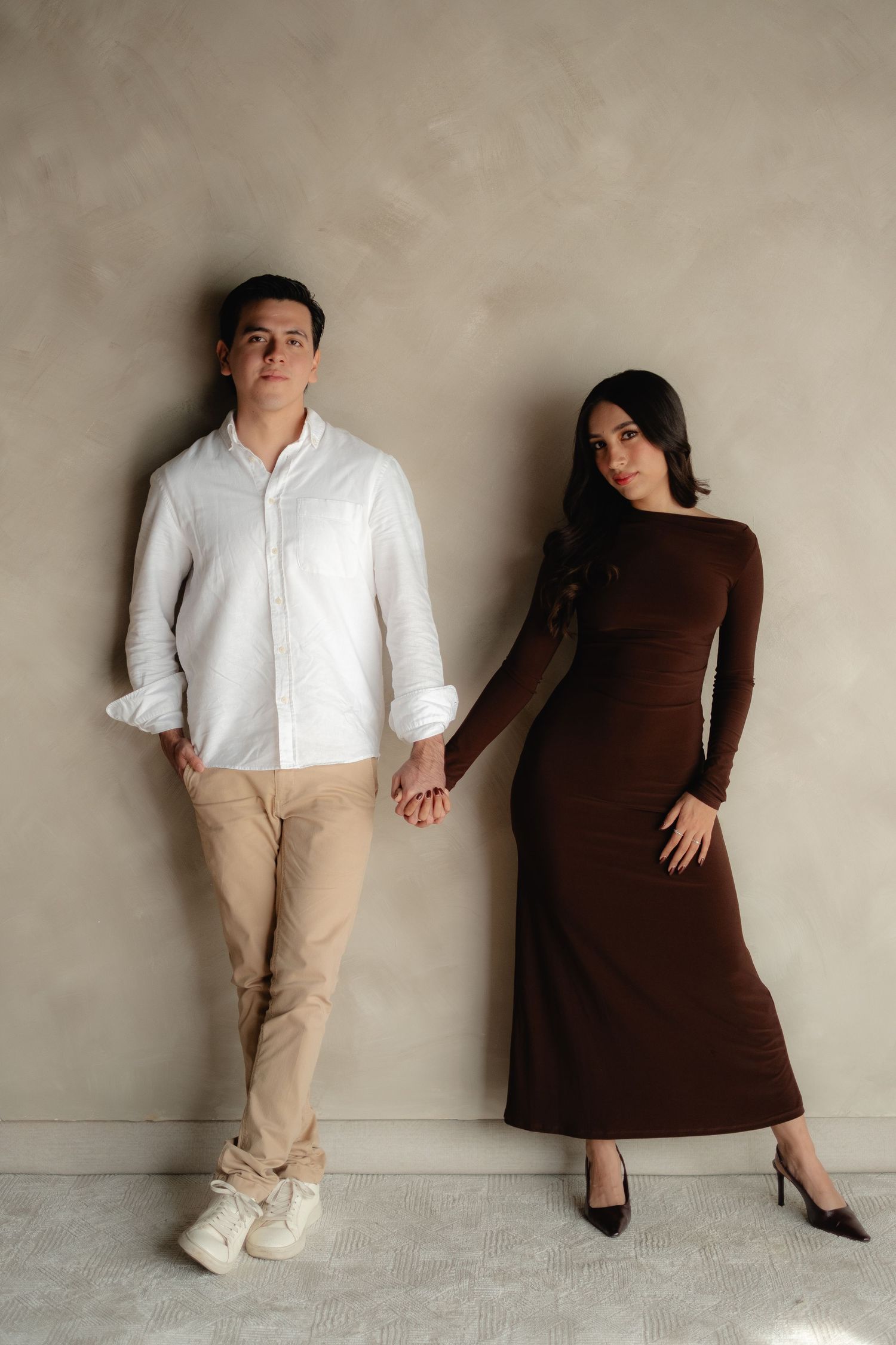Stylish couple holding hands against a gray wall, man in white shirt and woman in brown maxi dress.