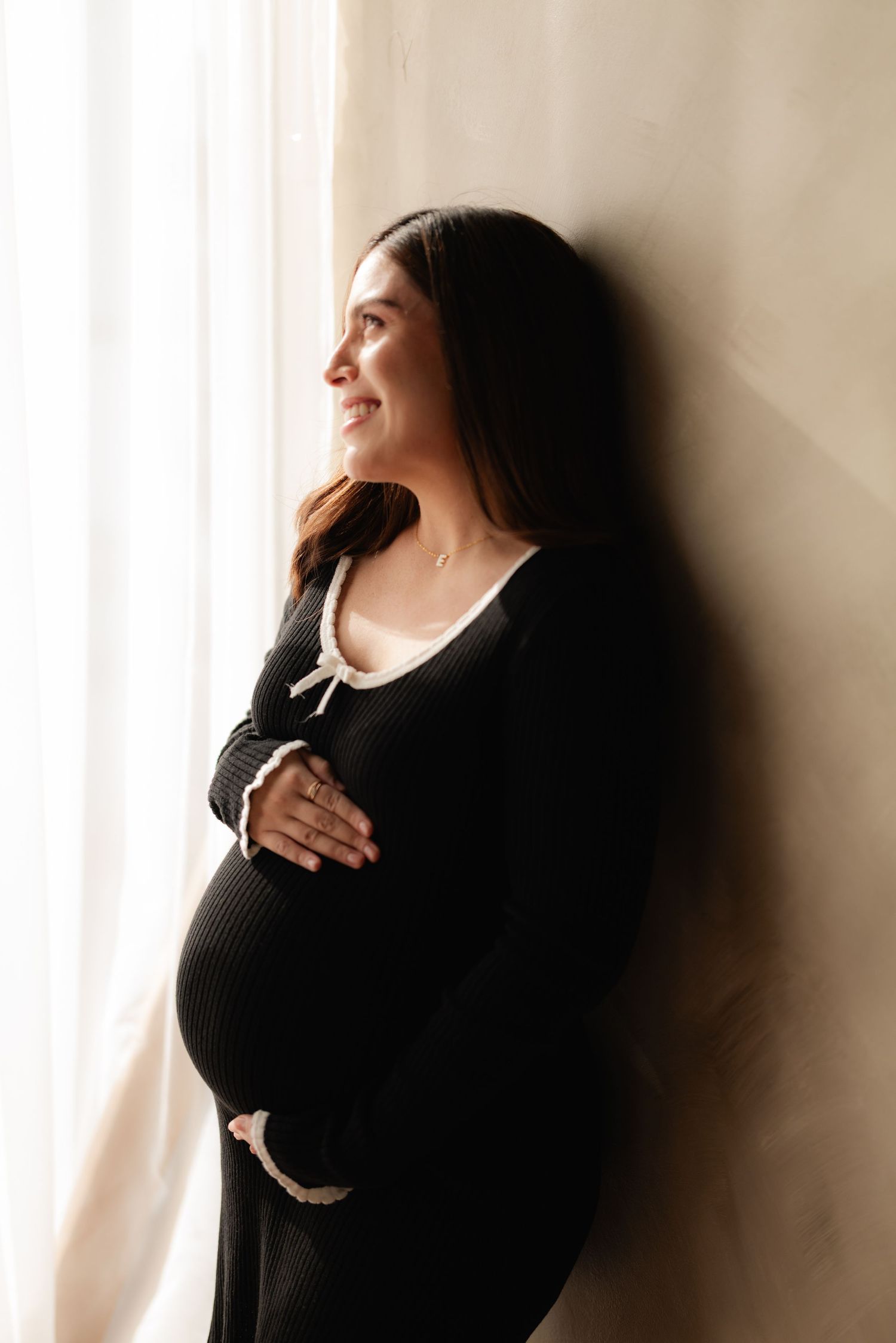 Pregnant woman in black ribbed dress leans against wall by window, smiling while cradling her baby bump.