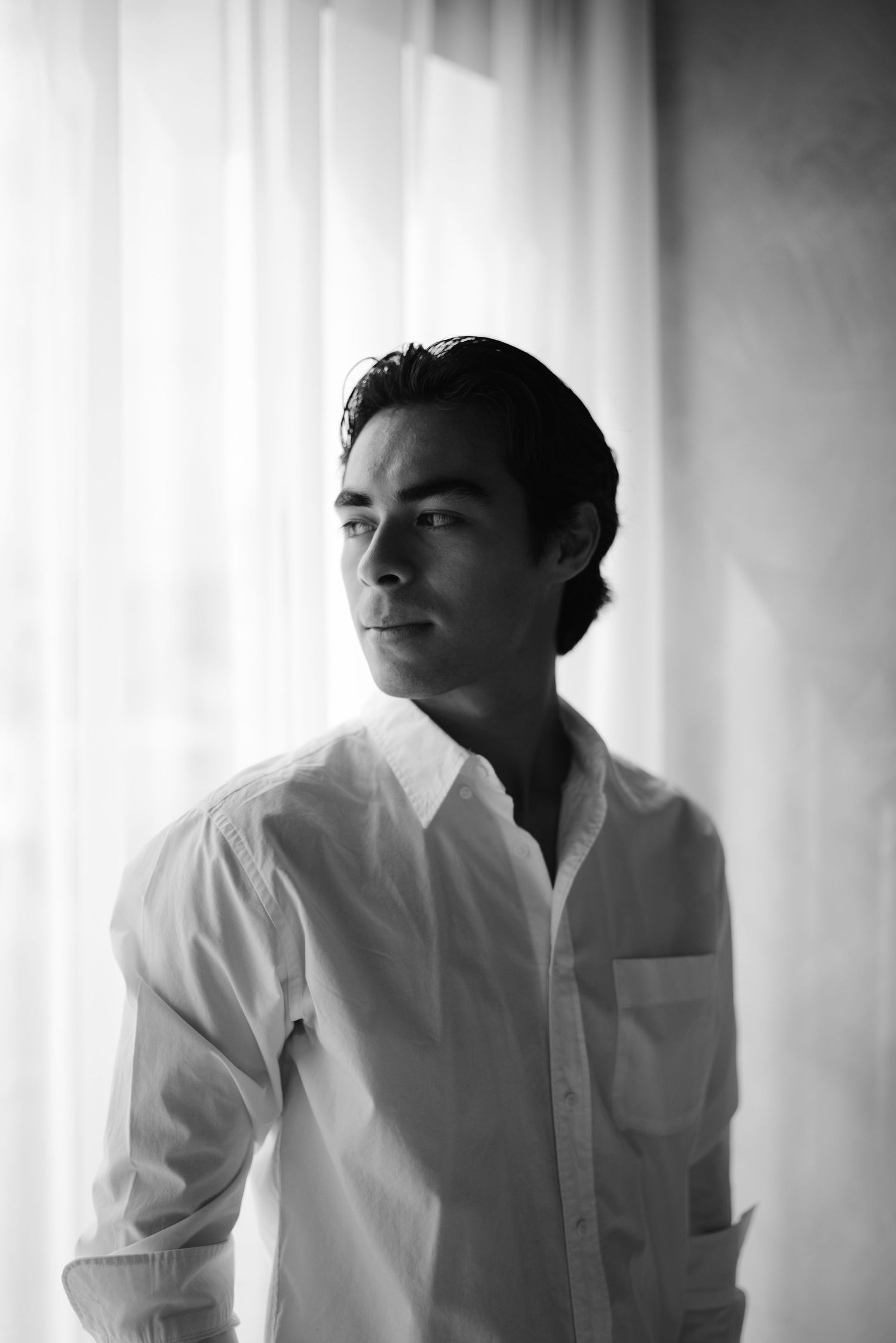 Black and white portrait of young man in white shirt gazing away by sheer curtains.
