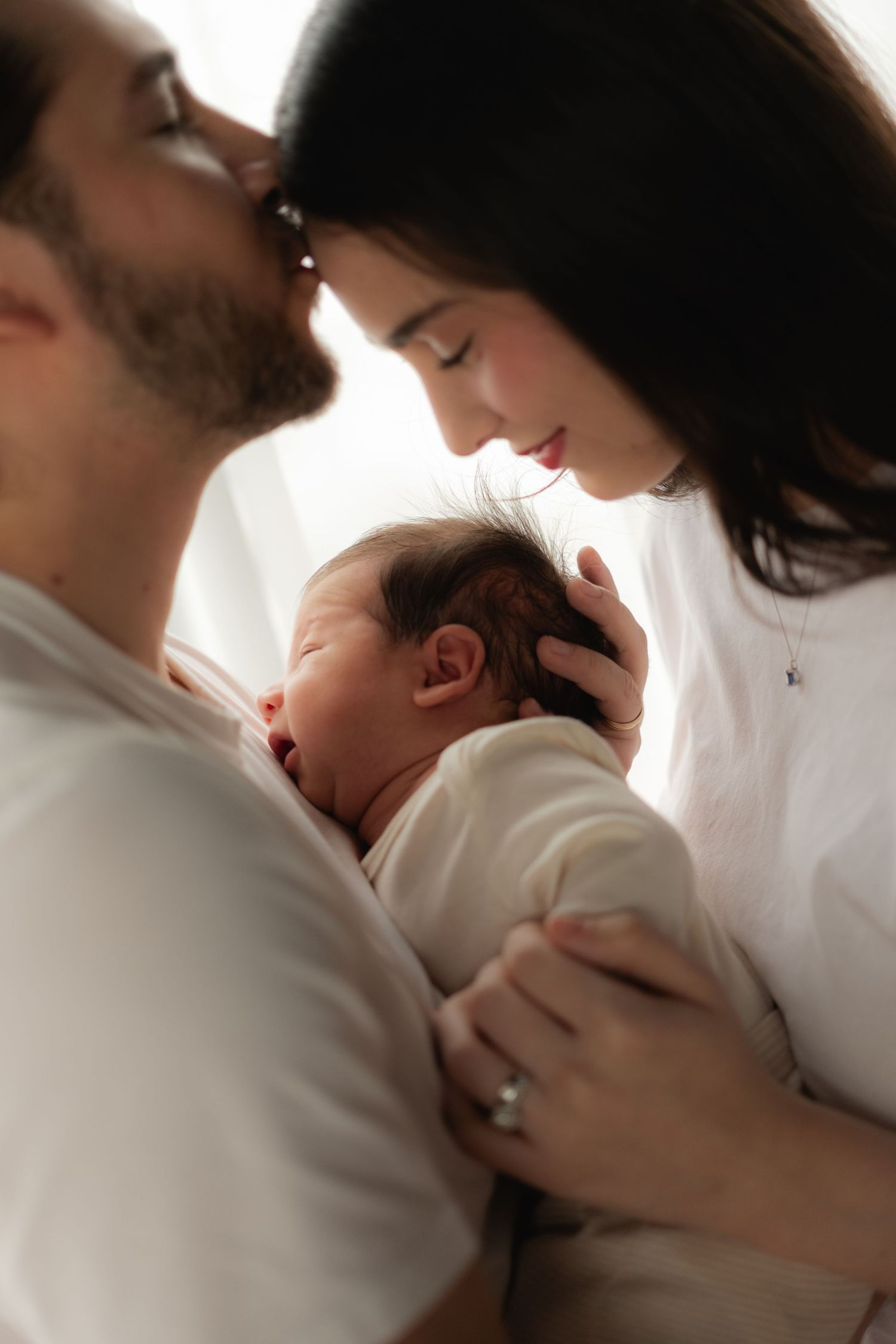 New parents lean in close together while gently cradling their sleeping newborn baby in white wrap.