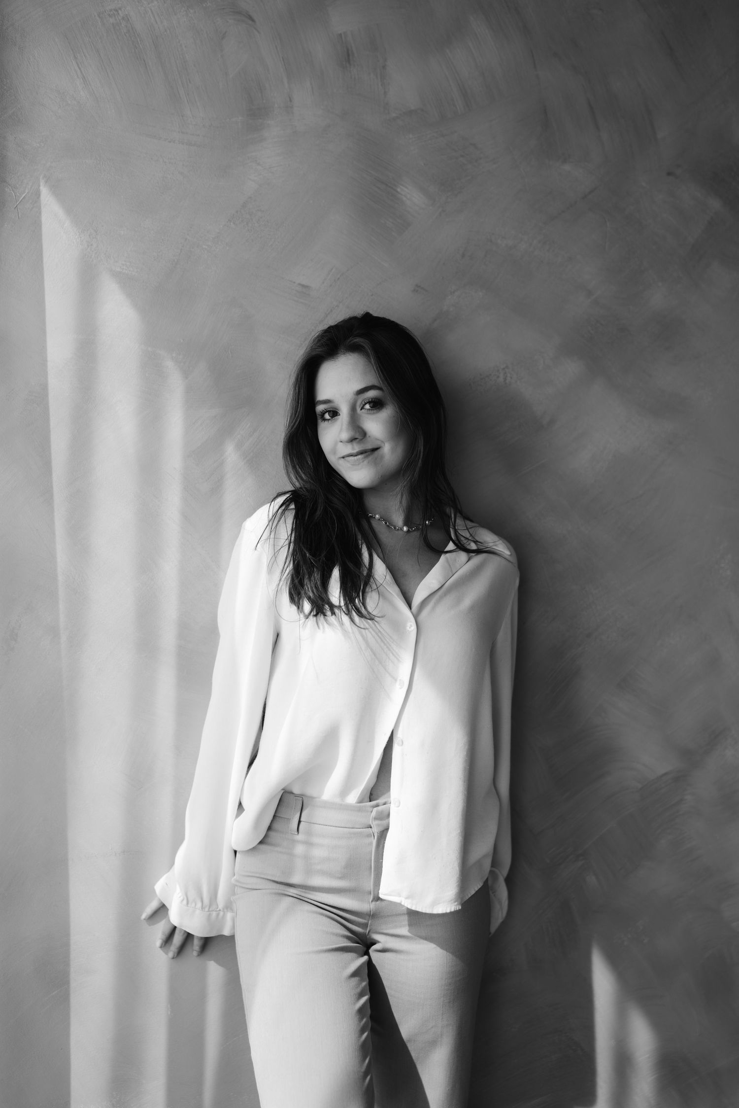 Black and white portrait of a young woman in a white blouse leaning against a textured wall.