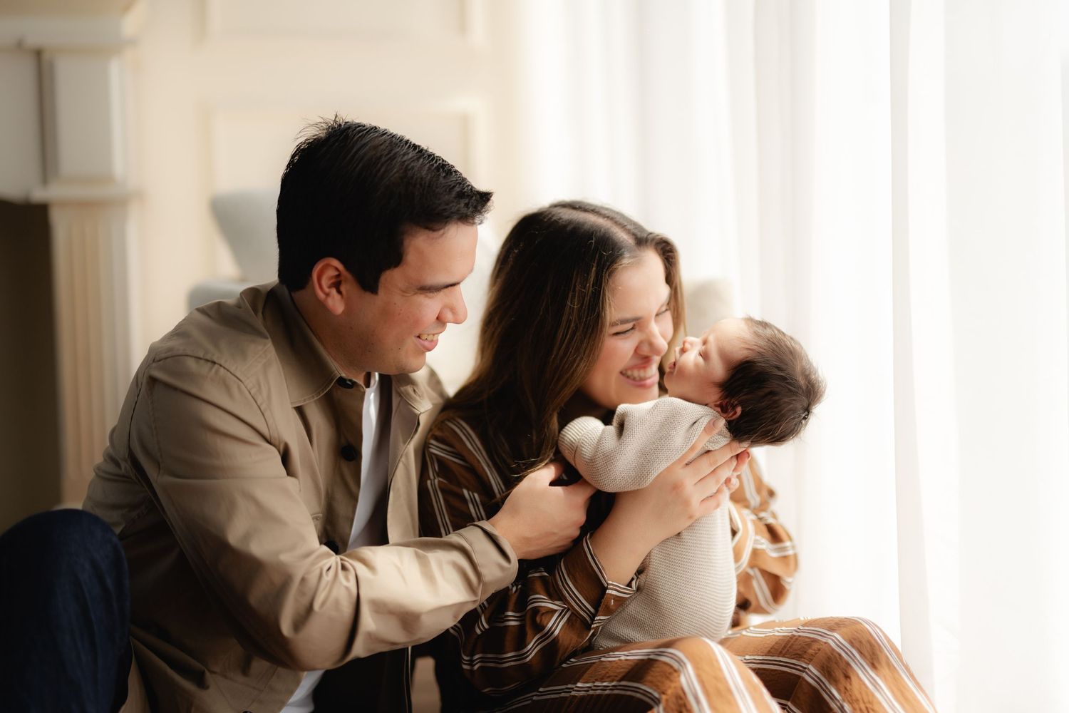 Happy young parents smiling at their newborn baby wrapped in white blanket near bright window.