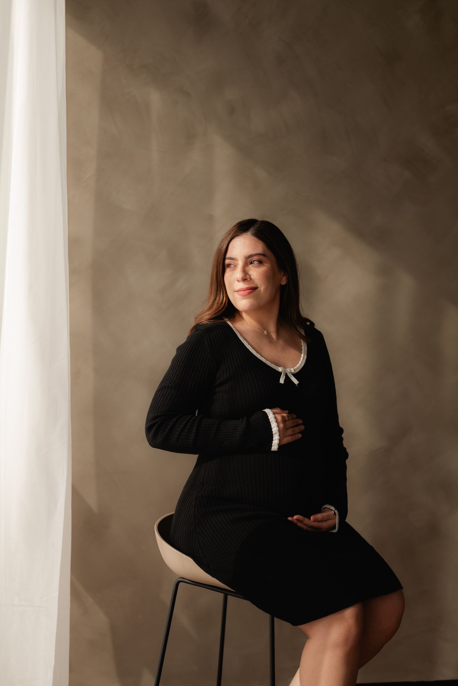 Elegant pregnant woman in black dress with white collar sits on stool, smiling softly in a studio setting.