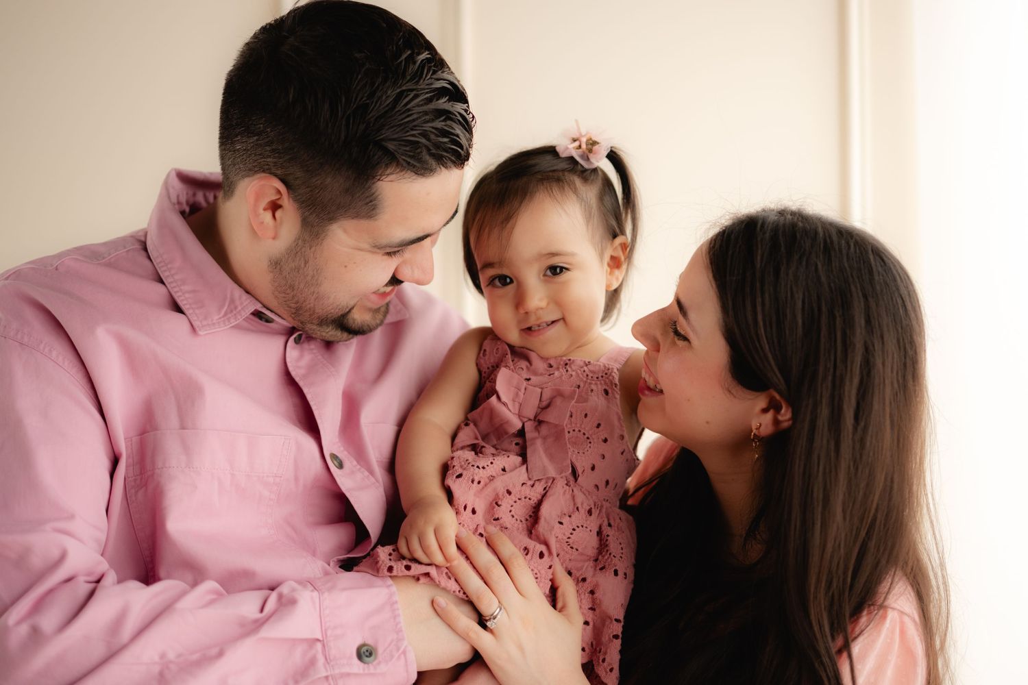 Happy young family with father in pink shirt holding smiling toddler girl beside mother indoors.