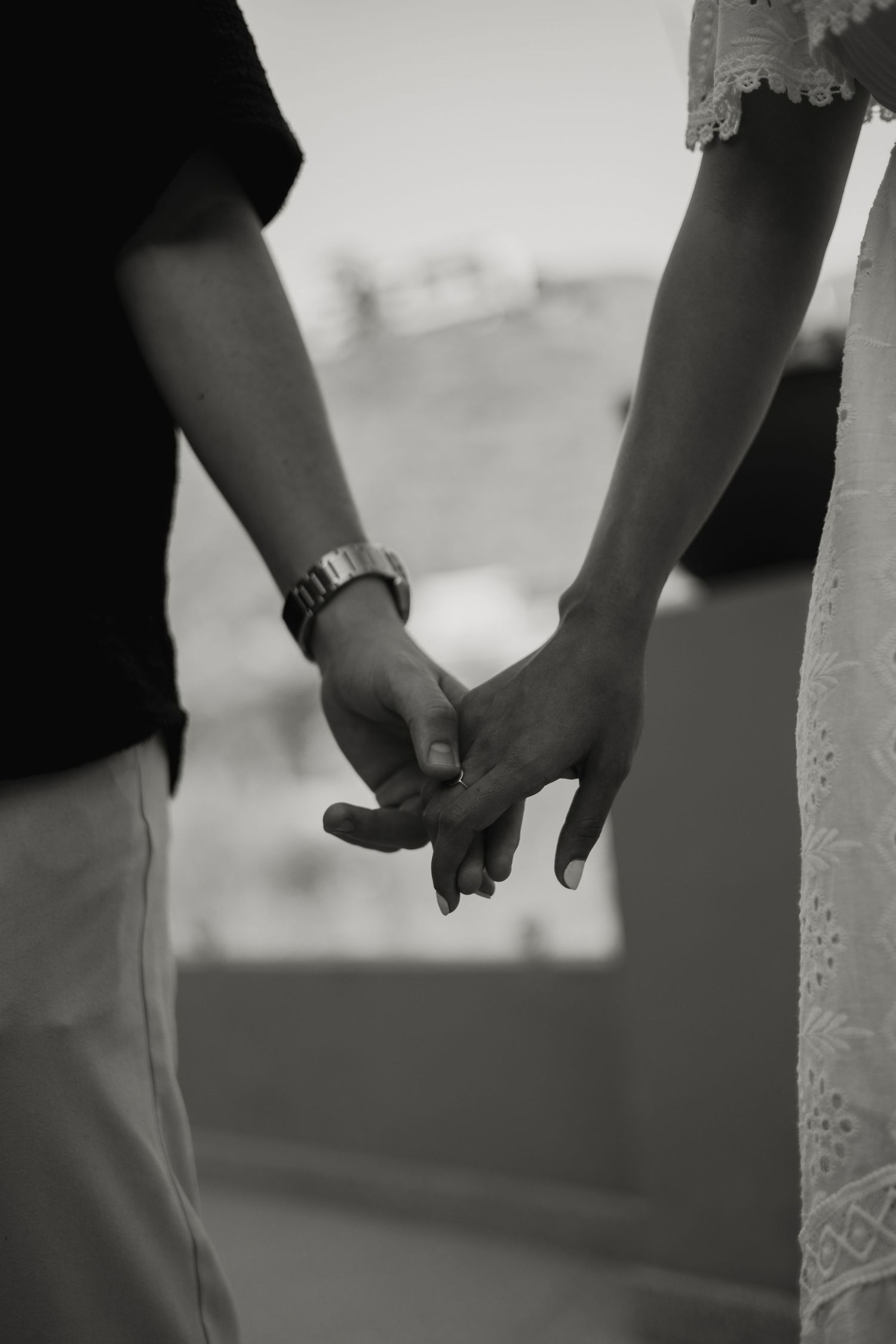 Black and white close-up of a couple holding hands, man wearing a watch, woman in lace dress.