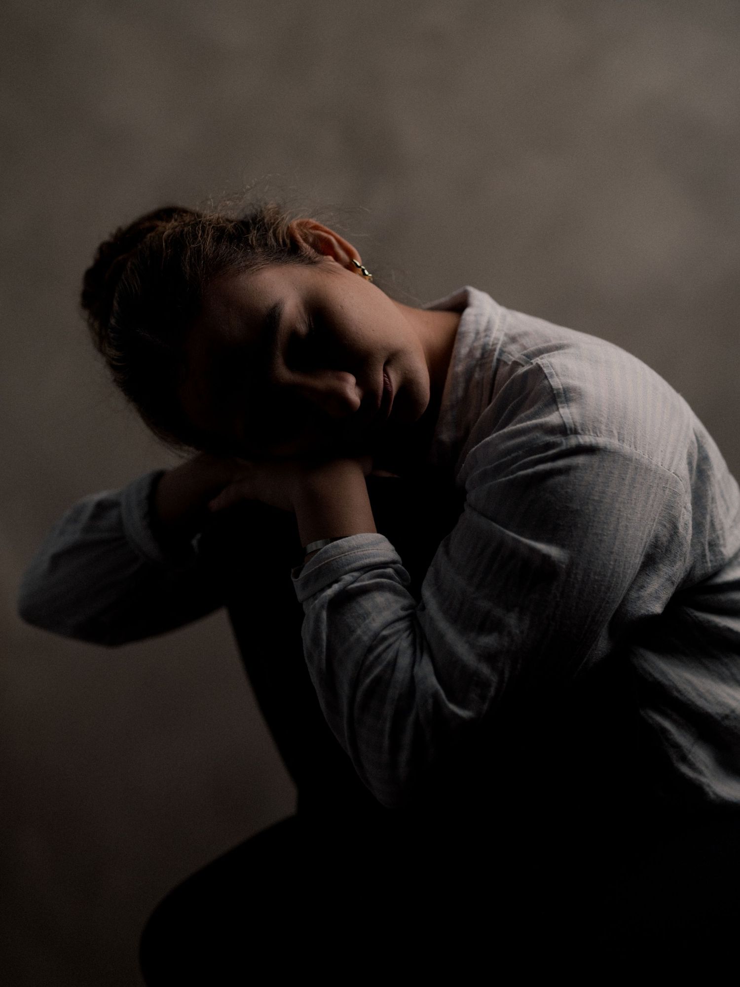 A young person sits in shadow, head resting on folded hands, conveying sadness or exhaustion in moody studio lighting.