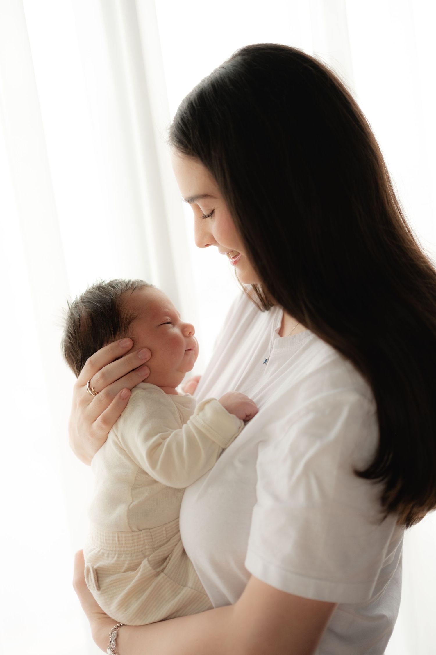 A mother tenderly holds her newborn baby, cradling its face while smiling softly in a bright, airy room.