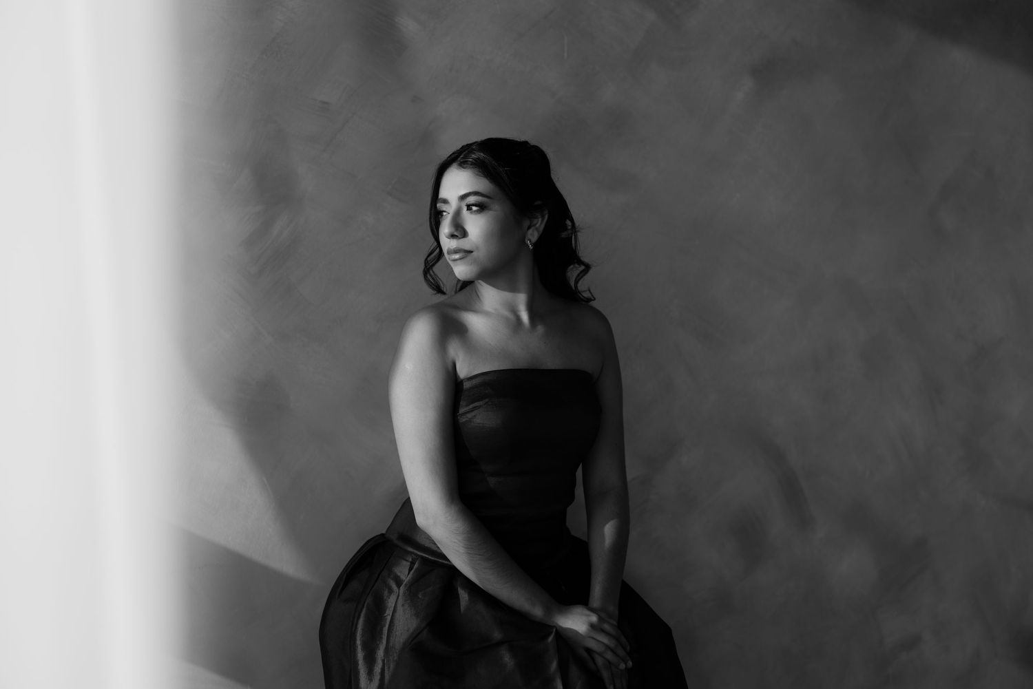 Elegant woman in strapless black gown sits pensively in a dramatic black and white studio portrait.