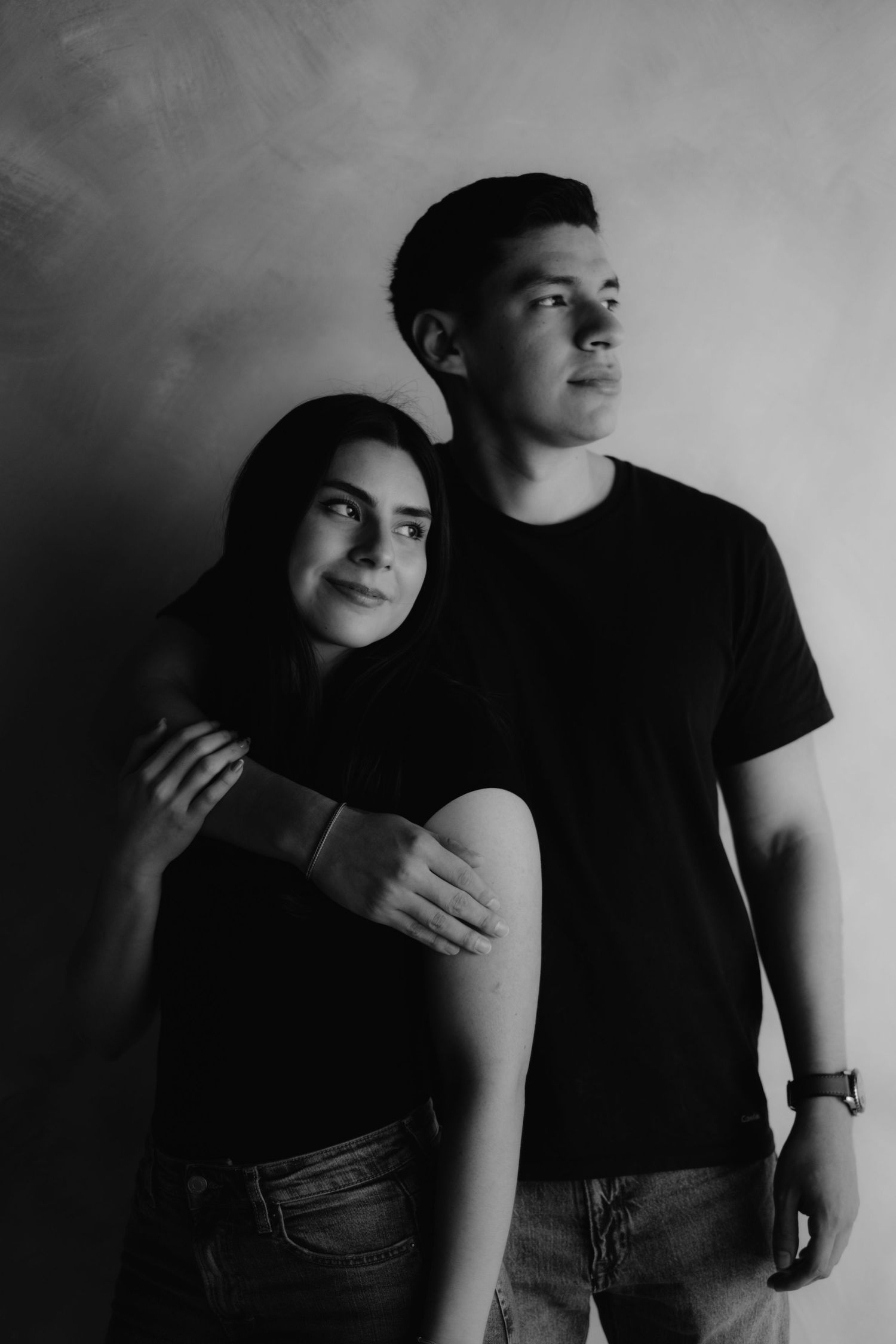 Black and white studio portrait of a young couple embracing against a textured gray wall.