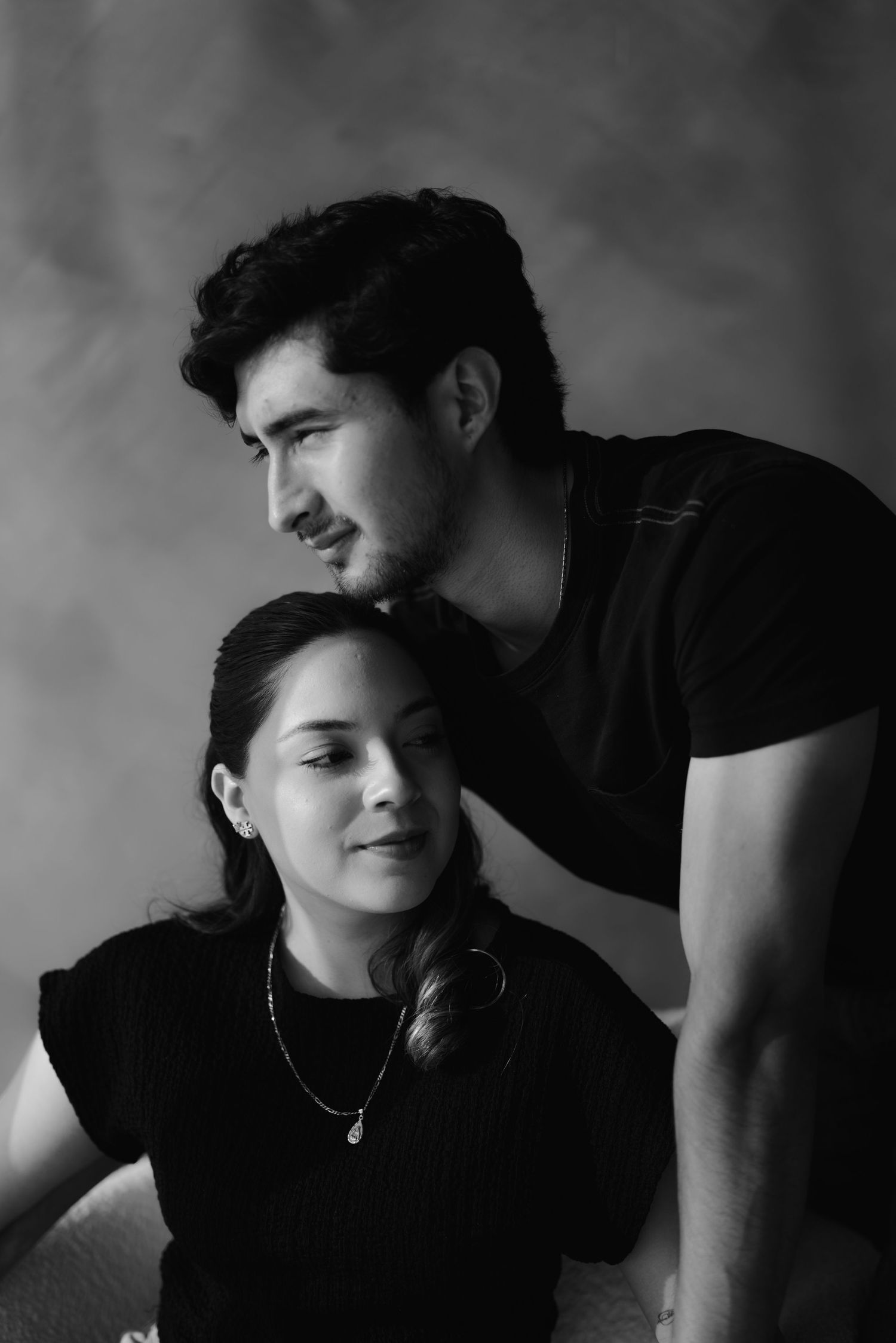 Black and white studio portrait of a young couple posing together against a textured gray background.