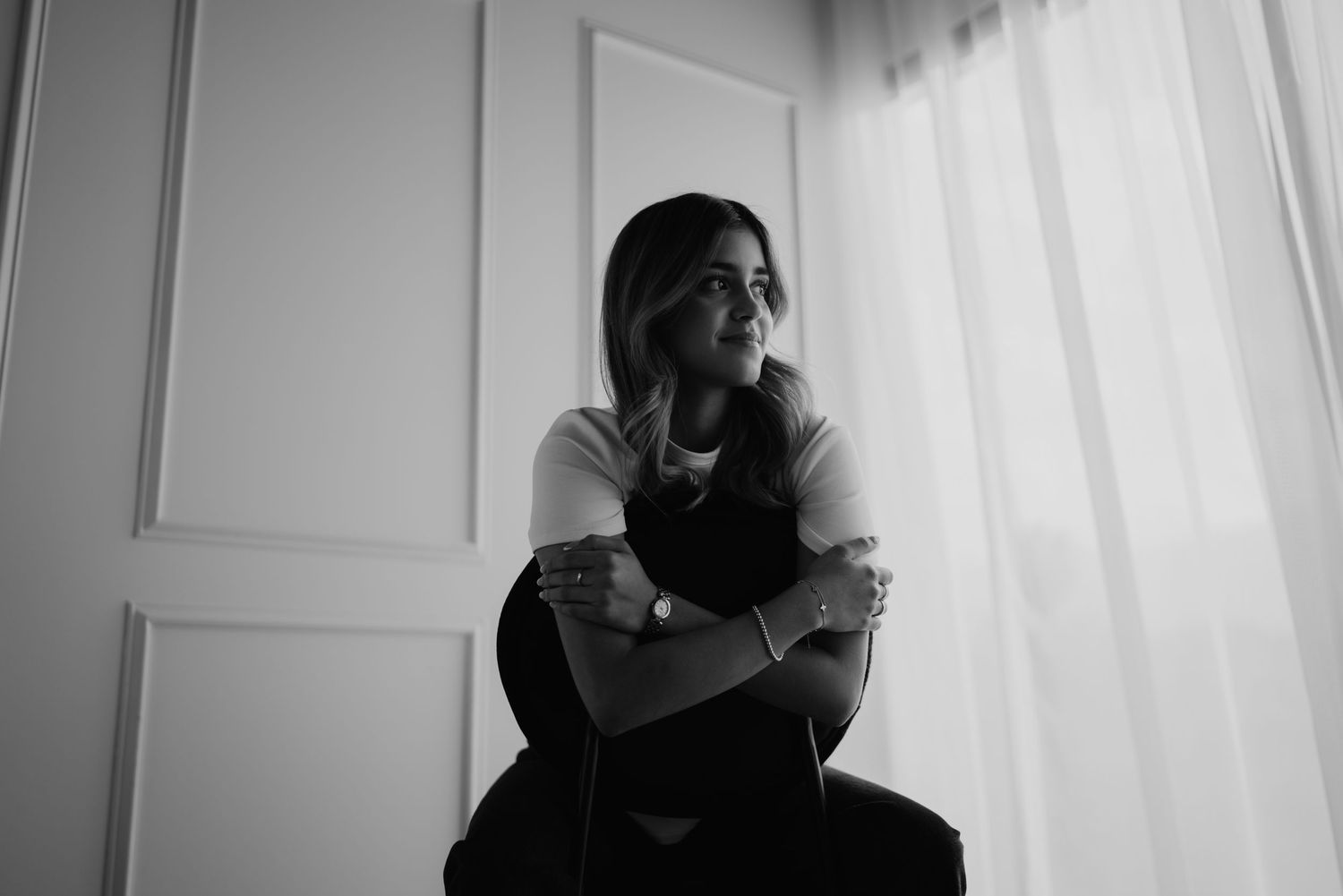Black and white photo of young woman sitting thoughtfully by bright window with arms crossed.