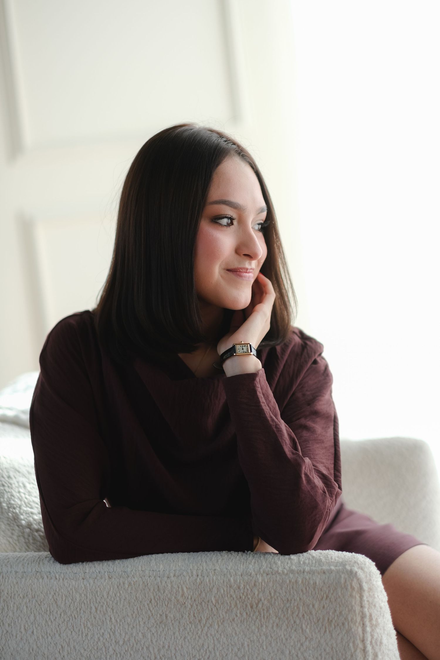 Pretty young woman with dark bob hair smiles warmly while relaxing in a bright, airy room.