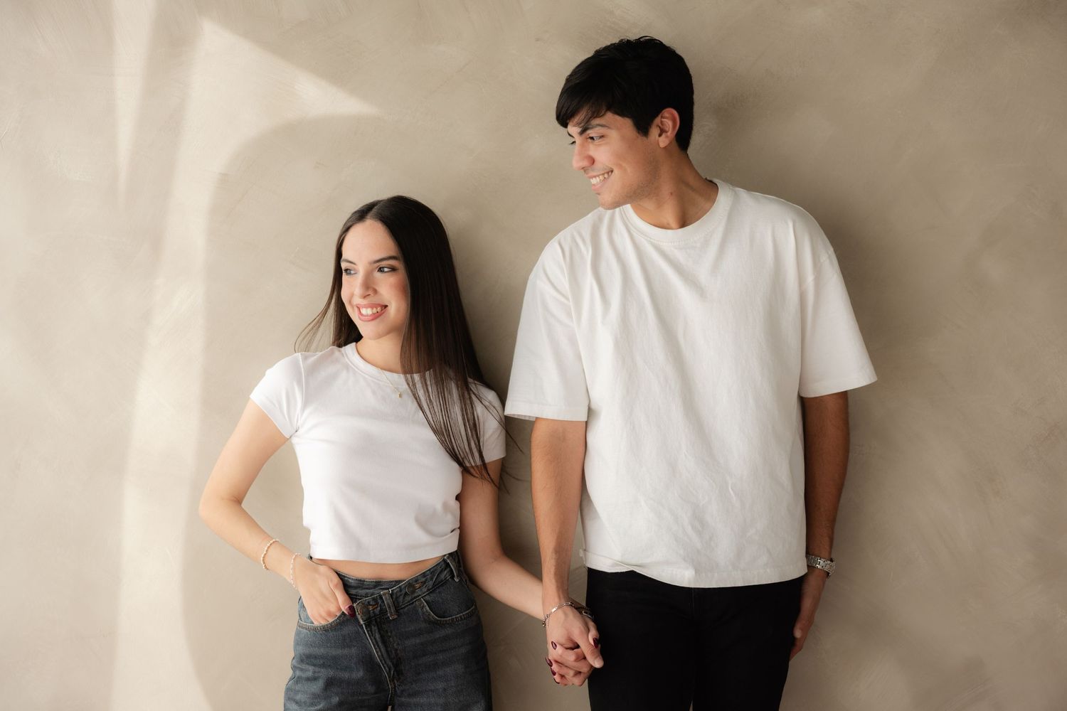 Young couple in matching white t-shirts holding hands and smiling against a beige wall.