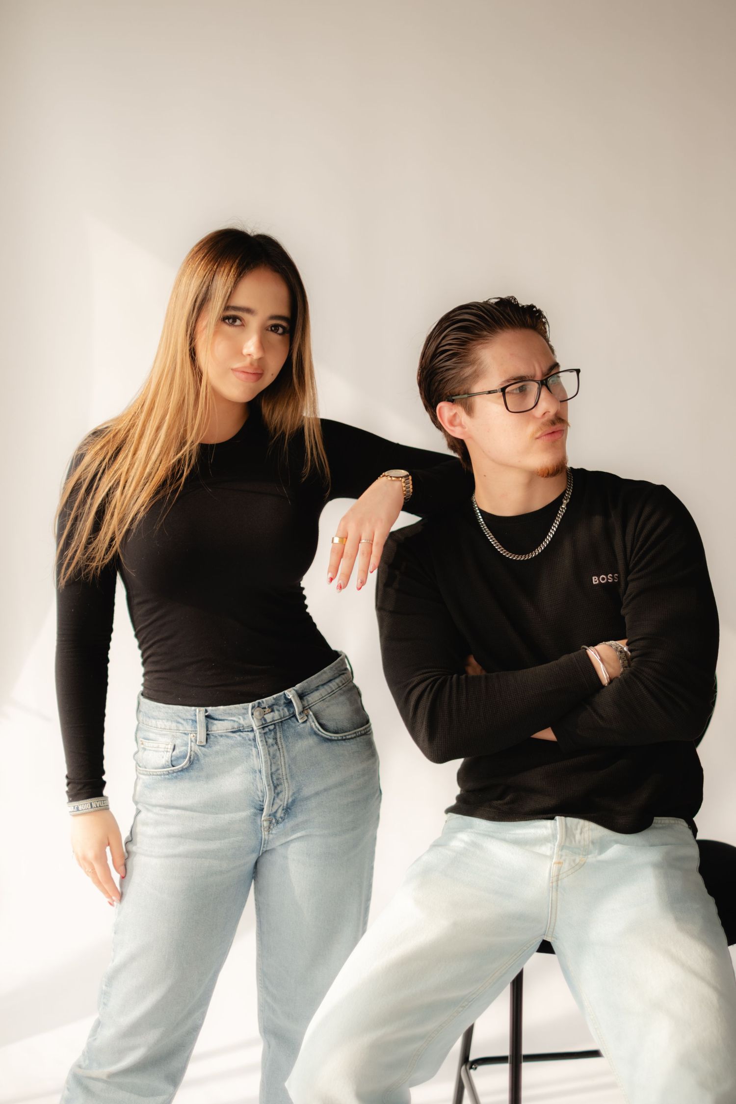 A young man and woman in matching black tops and light jeans pose together against a white background.