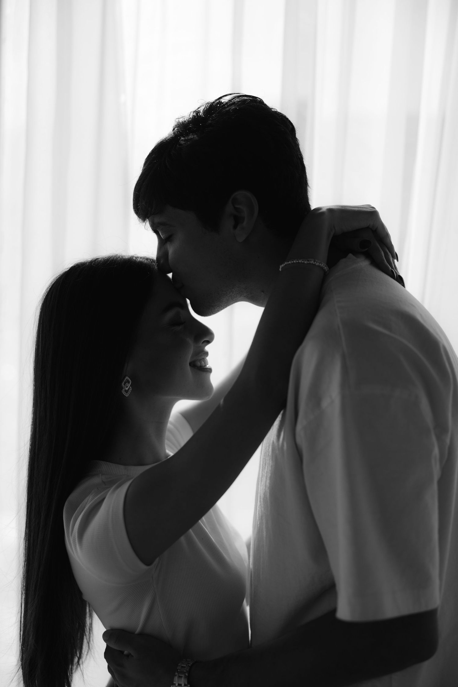 Black and white romantic couple portrait with backlit window glow, woman smiling up at man.