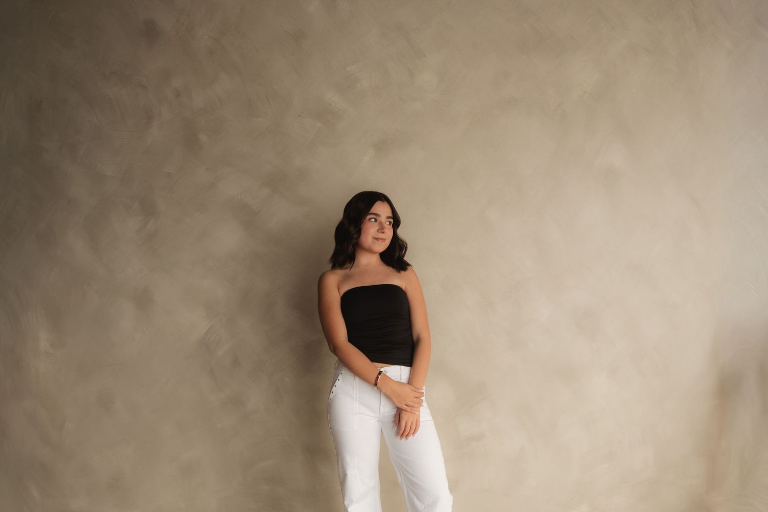Young woman in black tube top and white pants poses gracefully against a neutral textured wall.