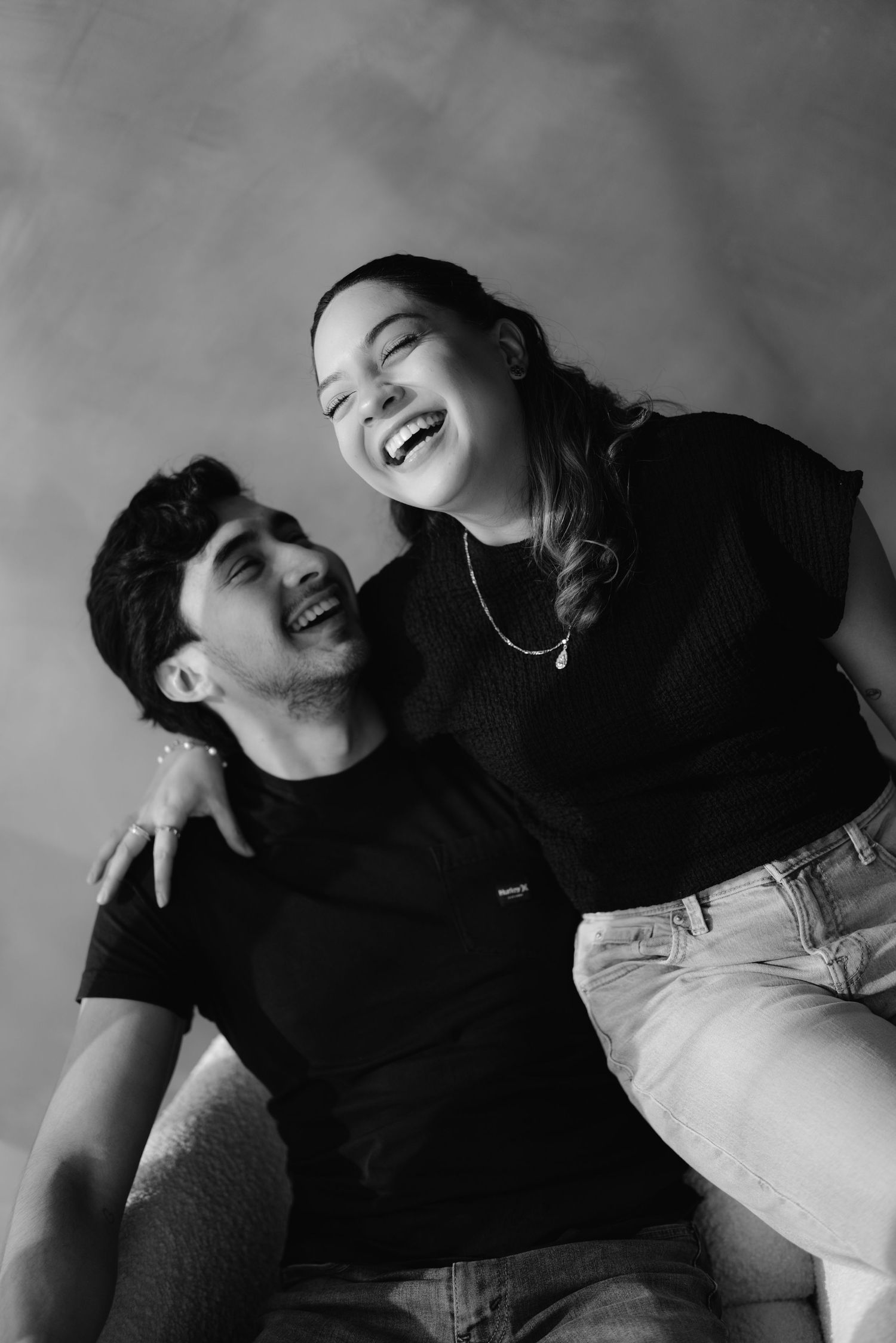 Black and white photo of a laughing young couple in casual black outfits posing together in a studio.