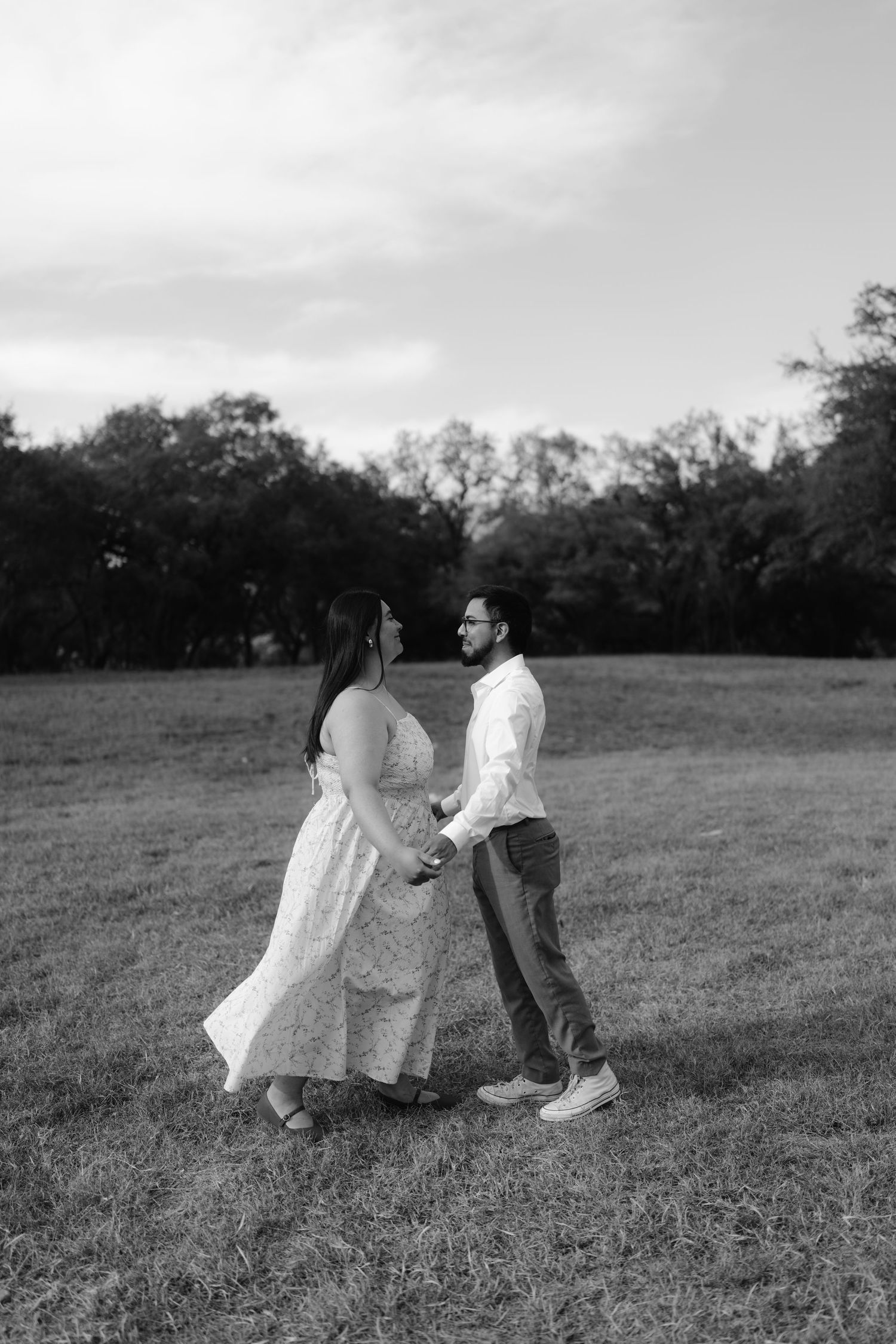 Black and white photo of a couple holding hands and gazing at each other in an open field with trees in the background.