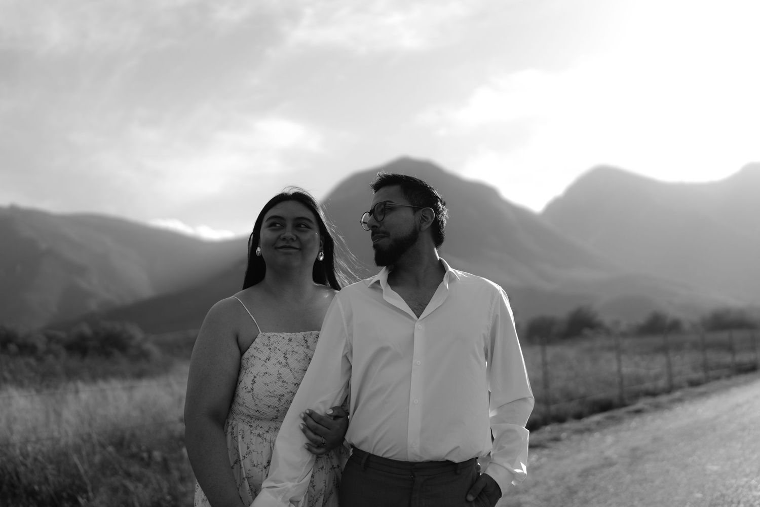 Black and white photo of a couple walking on a rural road with mountains in the background.