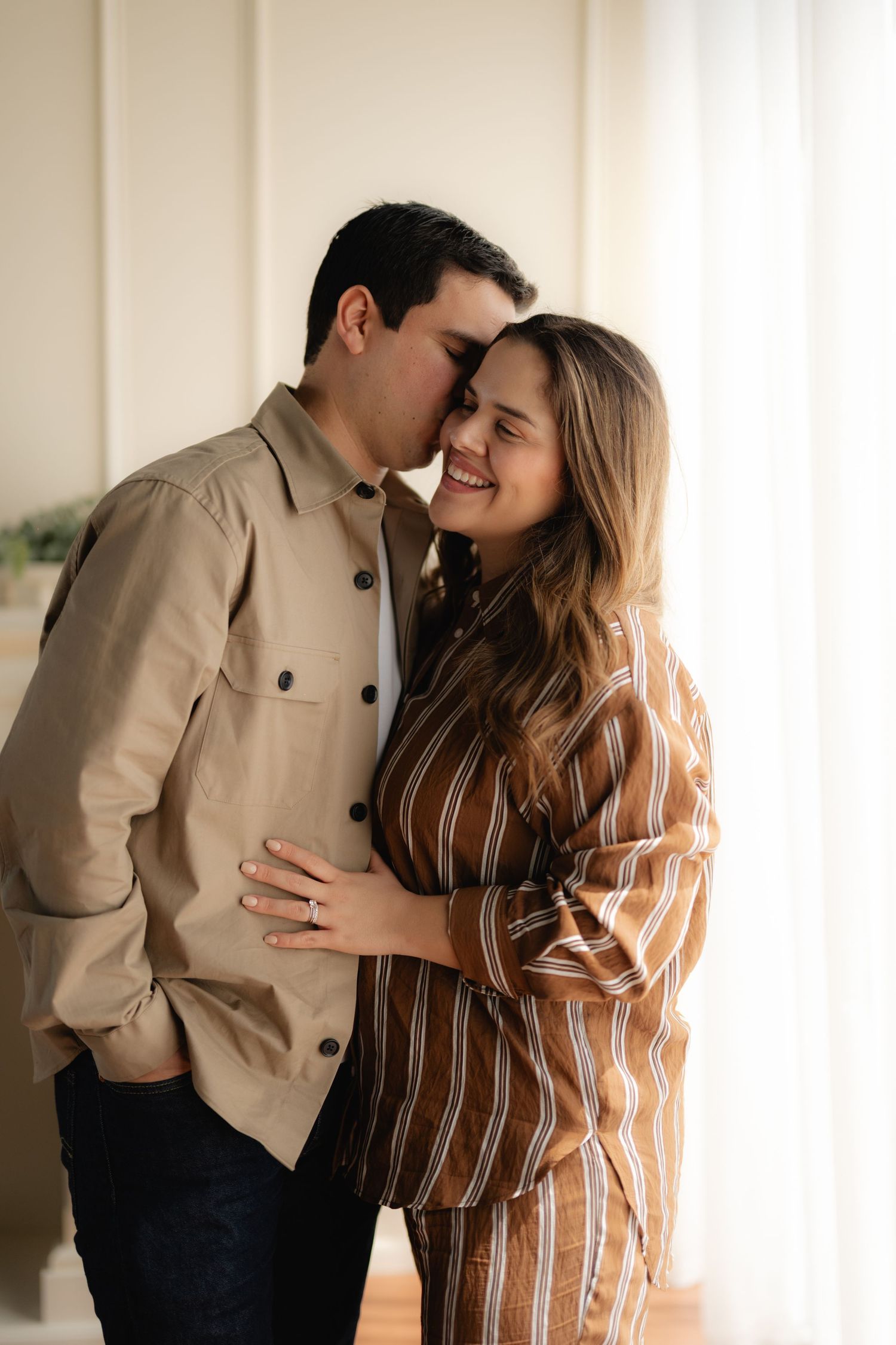 A couple shares a tender moment, the man kissing the smiling woman's temple as she embraces him in a bright, airy room.