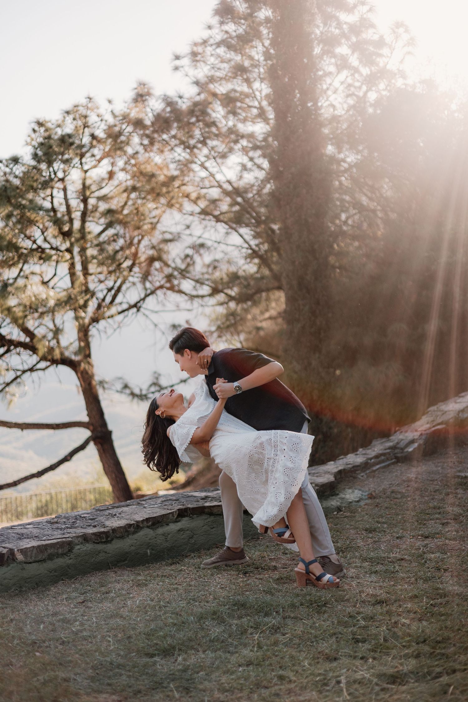 A couple shares a romantic dip kiss outdoors in golden sunlight surrounded by trees.