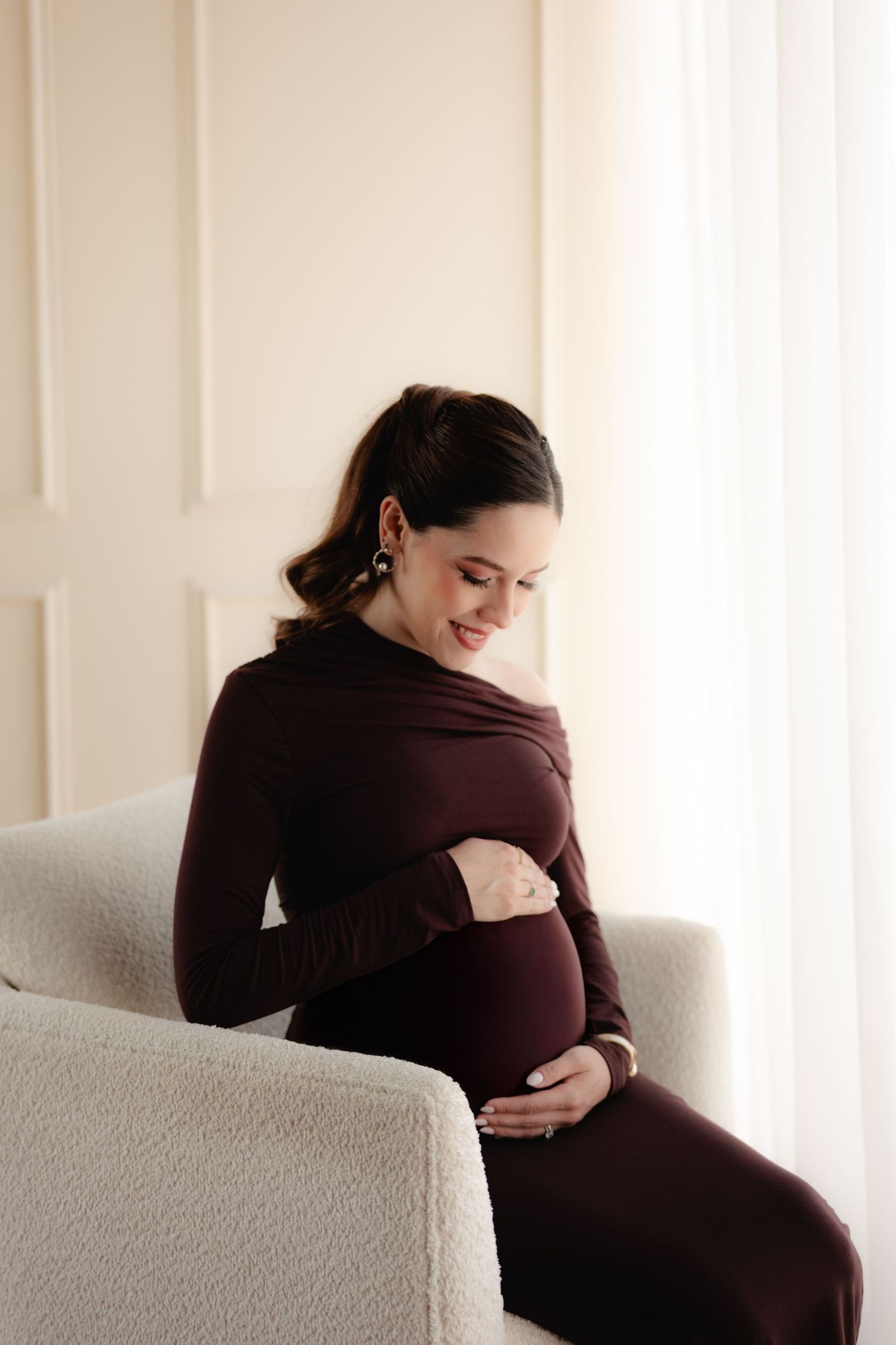 Pregnant woman in burgundy dress sitting in chair, smiling down at baby bump.