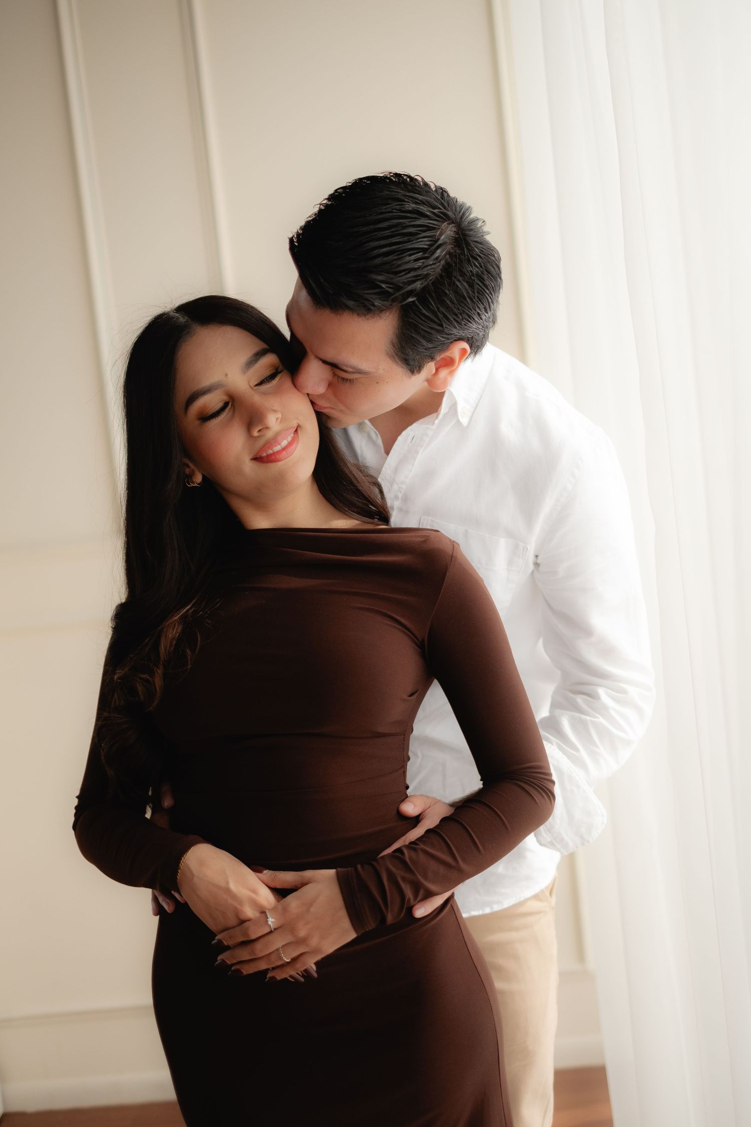 A man in a white shirt tenderly kisses a smiling woman in a brown dress near soft curtains.