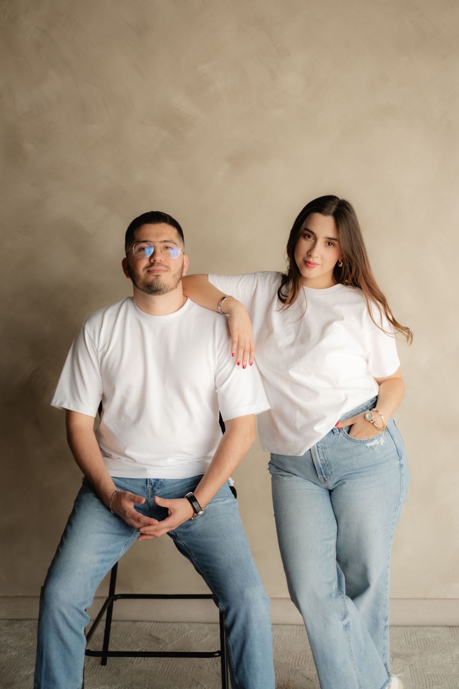 Couple in matching white t-shirts and jeans posing against beige wall, man seated on stool, woman standing beside him.