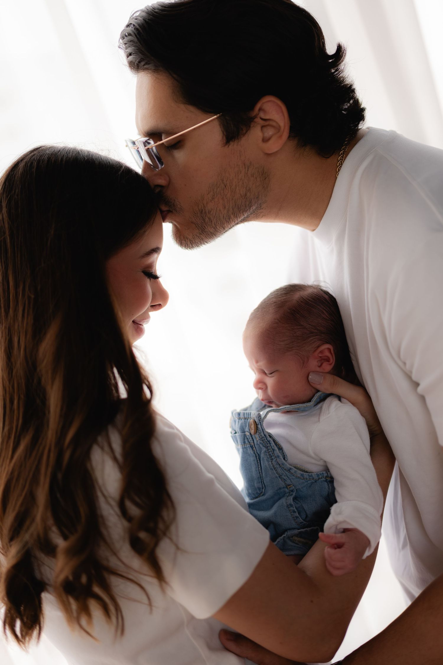 New parents in white shirts sharing tender kiss while mother holds newborn in denim overalls by window.