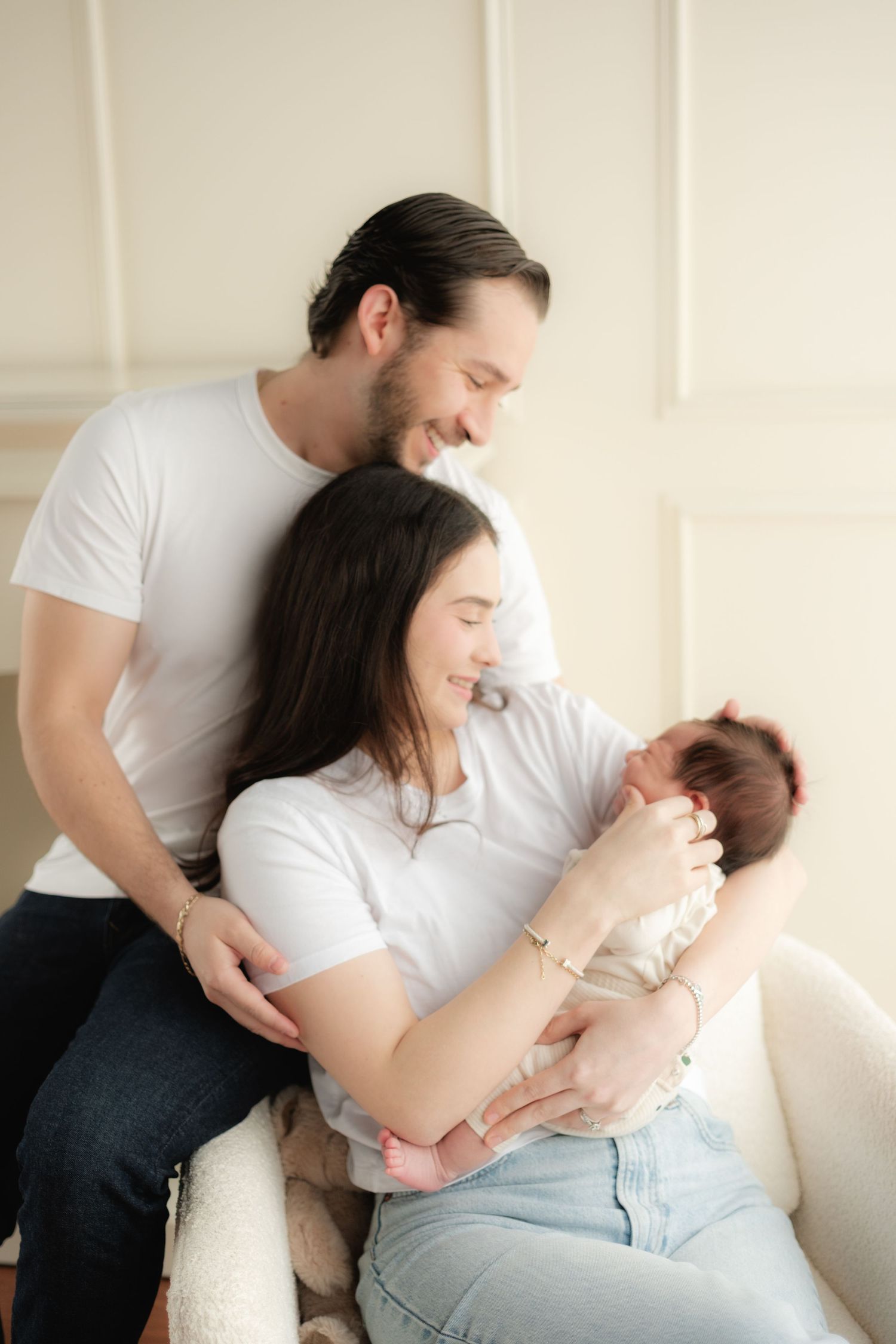 Happy new parents in white t-shirts smiling at newborn baby held by mother in cozy bright room.