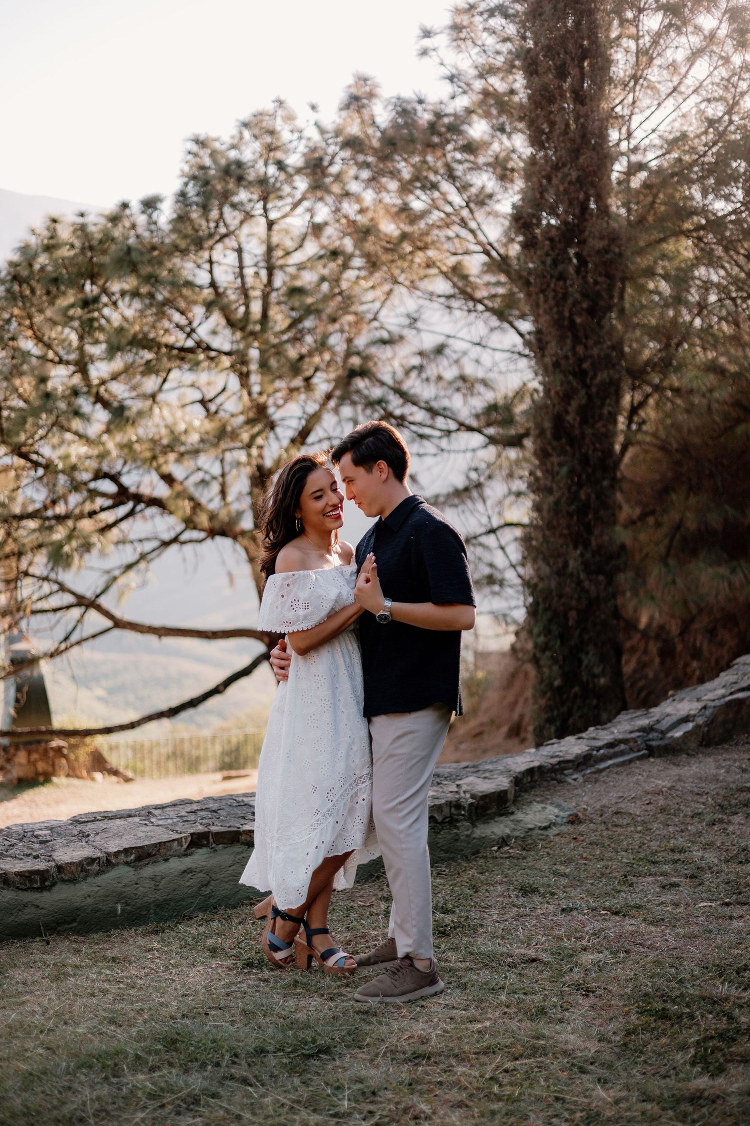 Couple dancing and smiling together outdoors at golden hour beneath a large tree in nature.