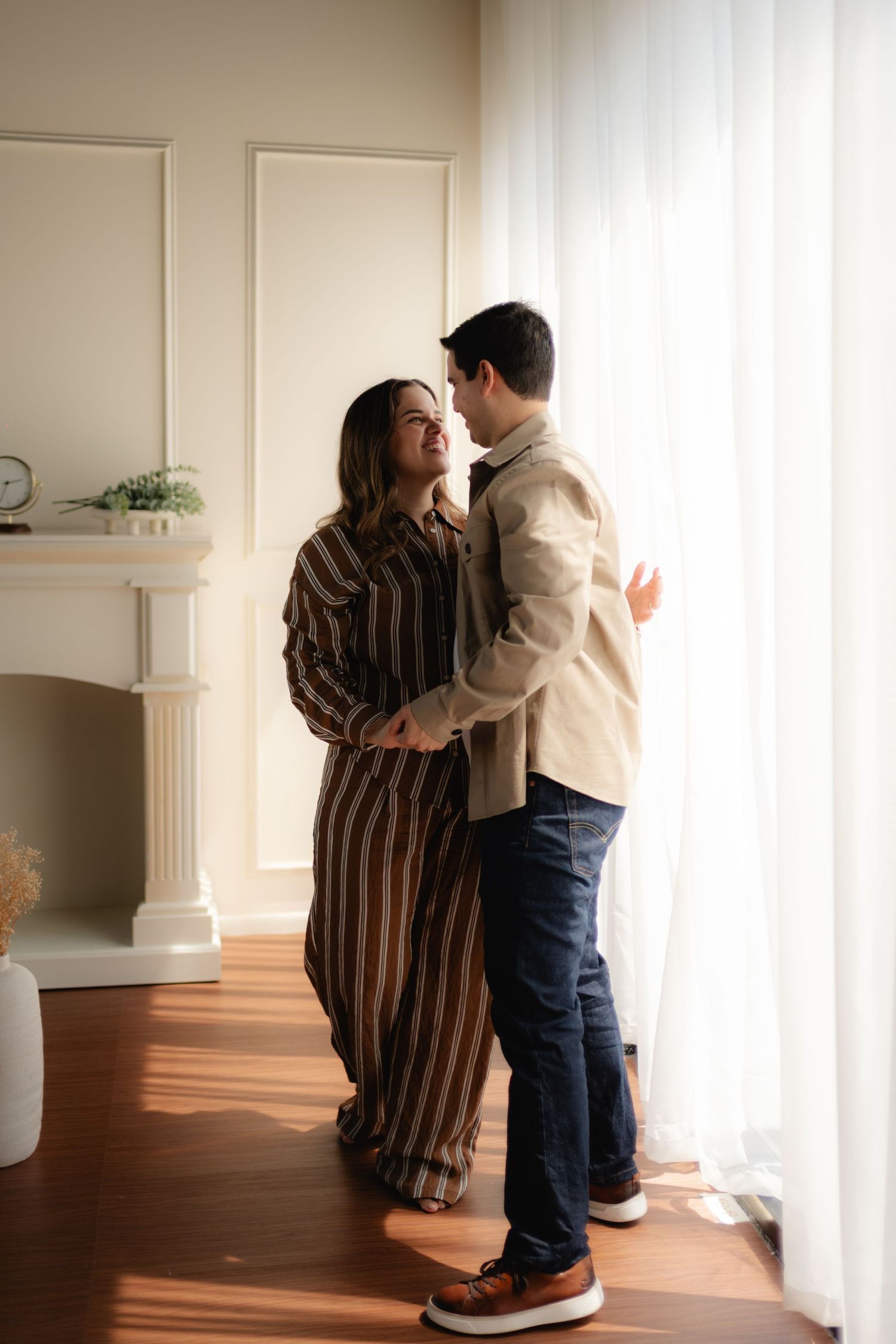 A couple embraces near a sunlit window in a cozy living room, wearing neutral tones.