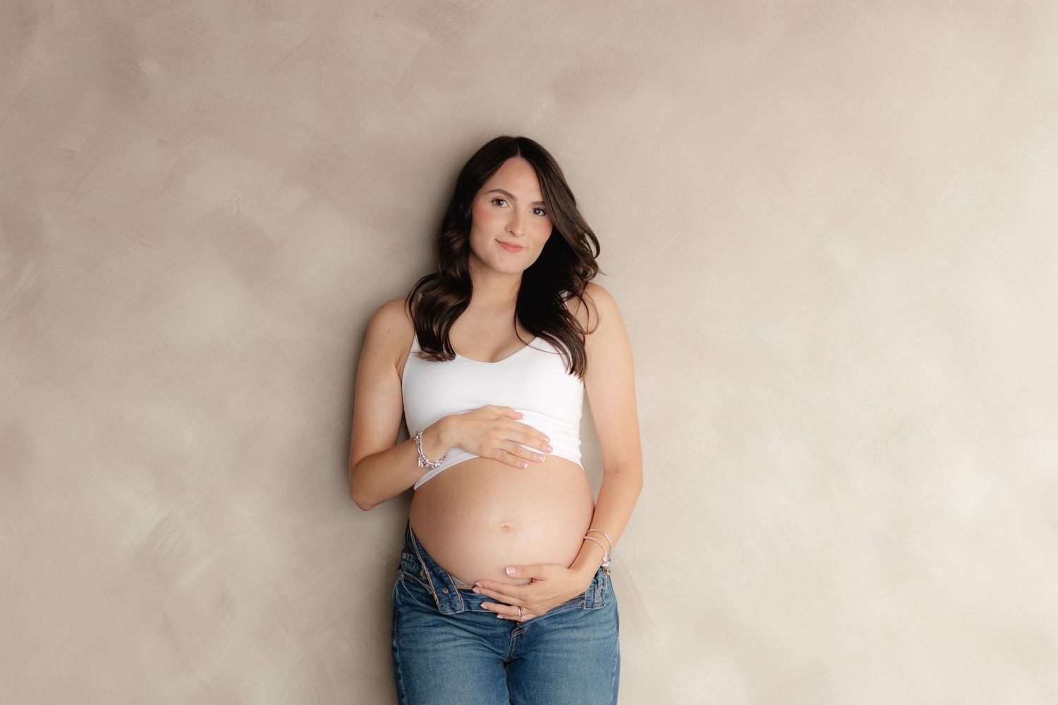 Pregnant woman in white crop top and jeans cradling her baby bump against a neutral beige studio backdrop.