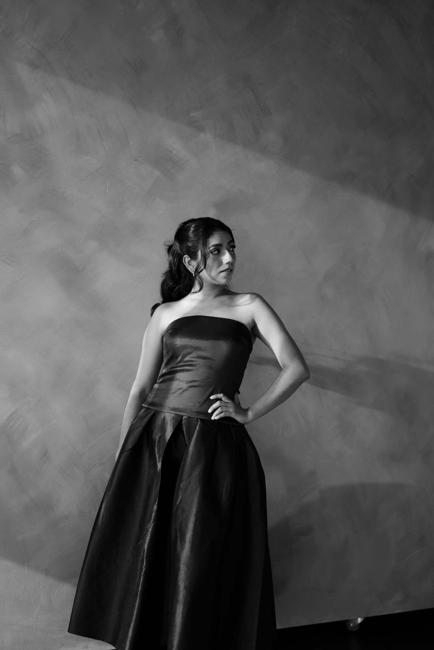 A woman in a strapless black ball gown poses confidently against a textured gray studio backdrop.