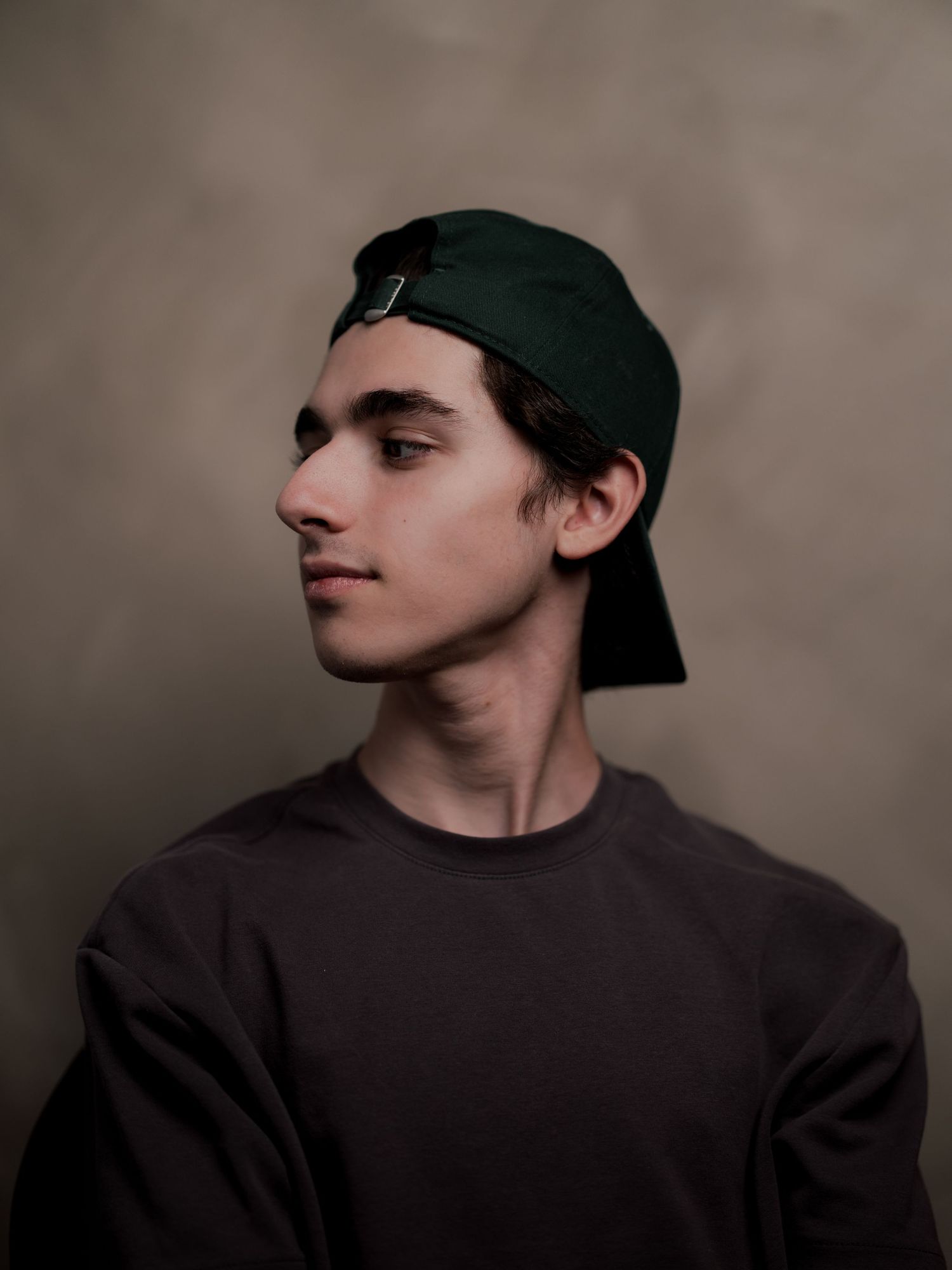 Young man wearing a dark green snapback cap and gray sweatshirt, posing in profile against a neutral background.