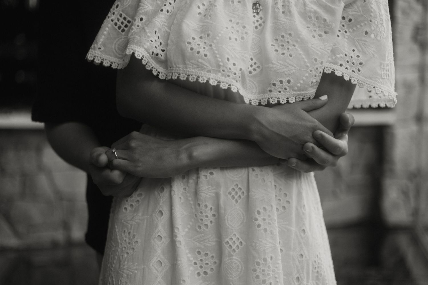 Black and white photo of a couple embracing, woman in white lace off-shoulder dress, hands intertwined romantically.