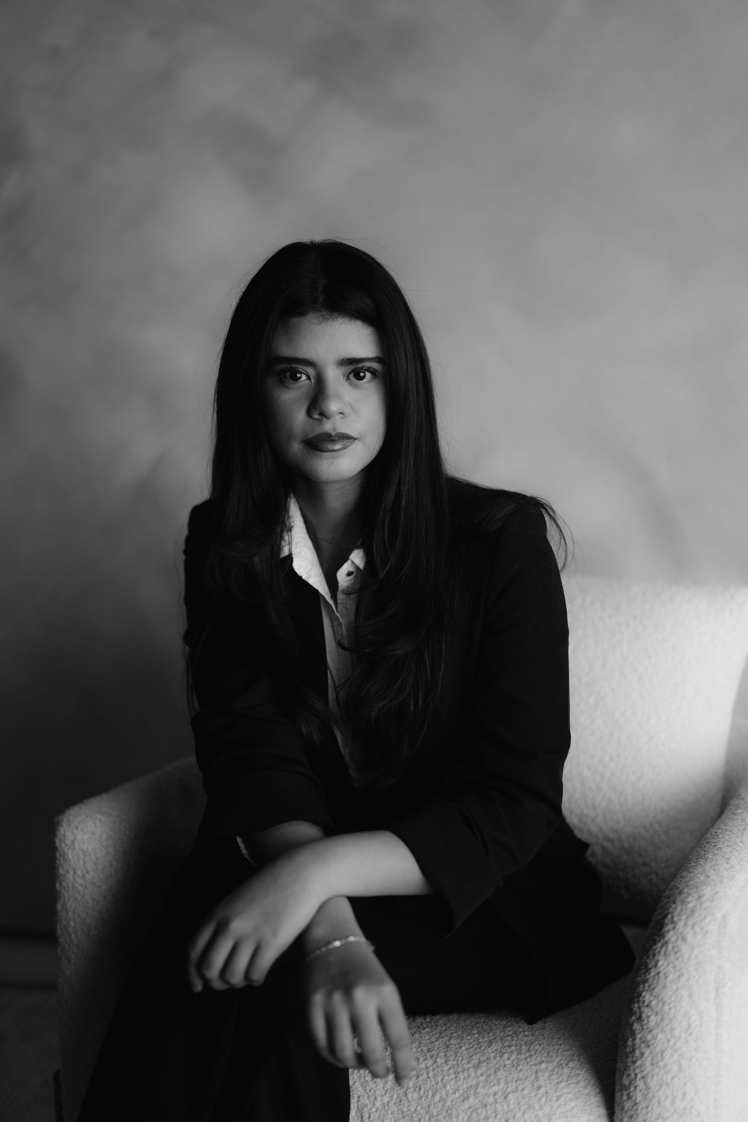 Young woman with long dark hair in black blazer seated in white chair, moody B&W portrait.