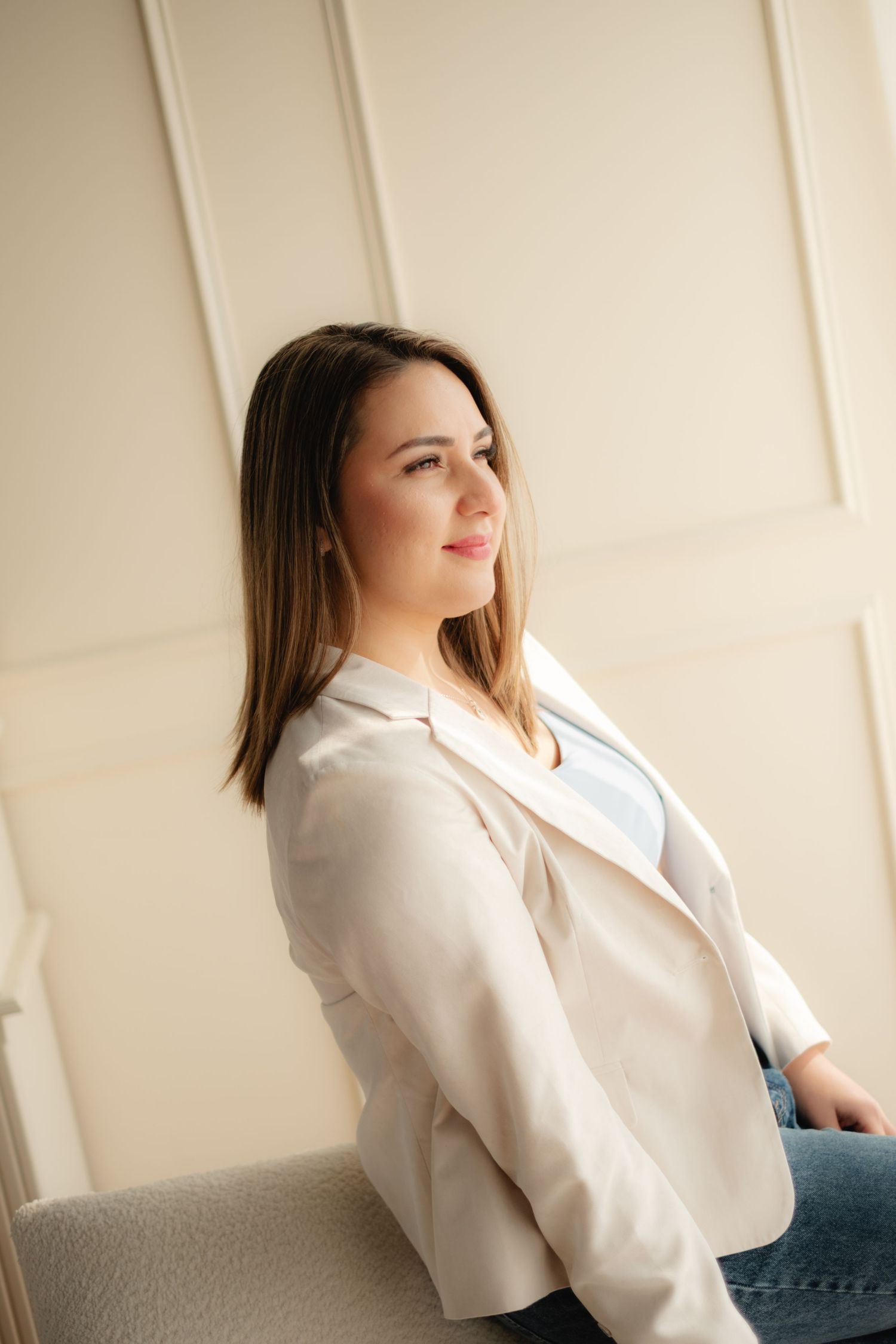 Elegant young woman in cream blazer sits relaxed and smiling in a bright, softly lit studio setting.