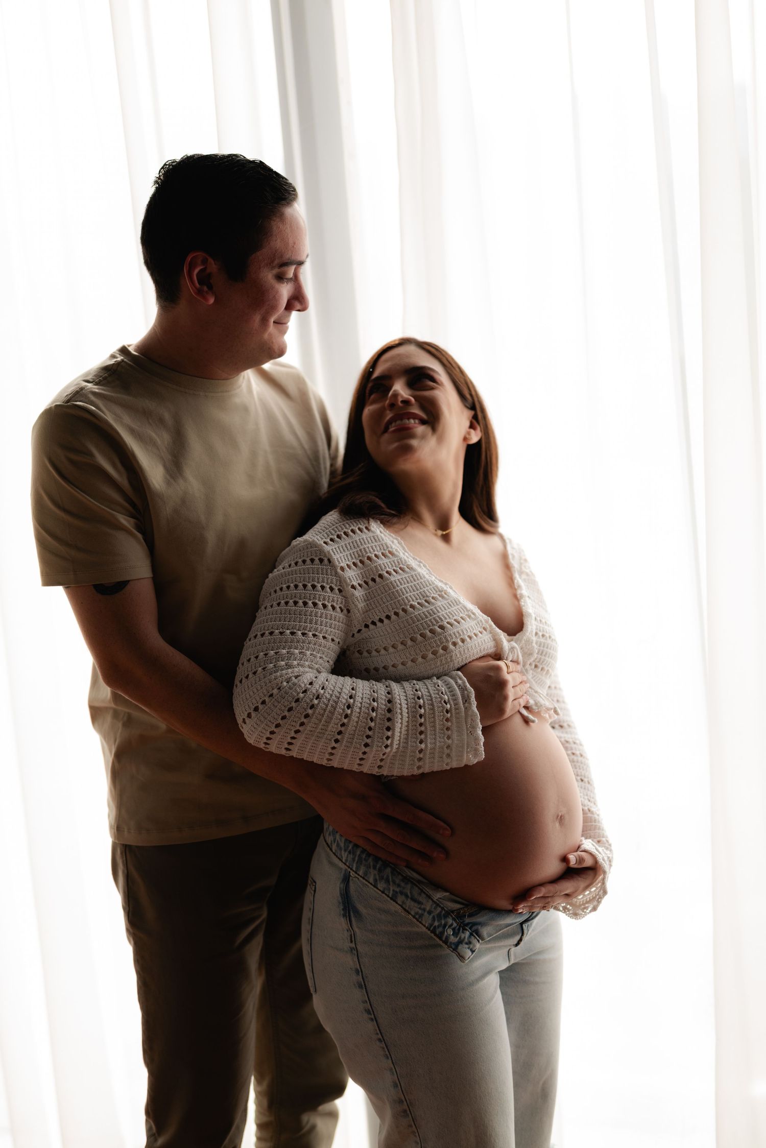 Couple embracing near sheer curtains, man hugging pregnant woman from behind as she smiles and holds her bare belly.