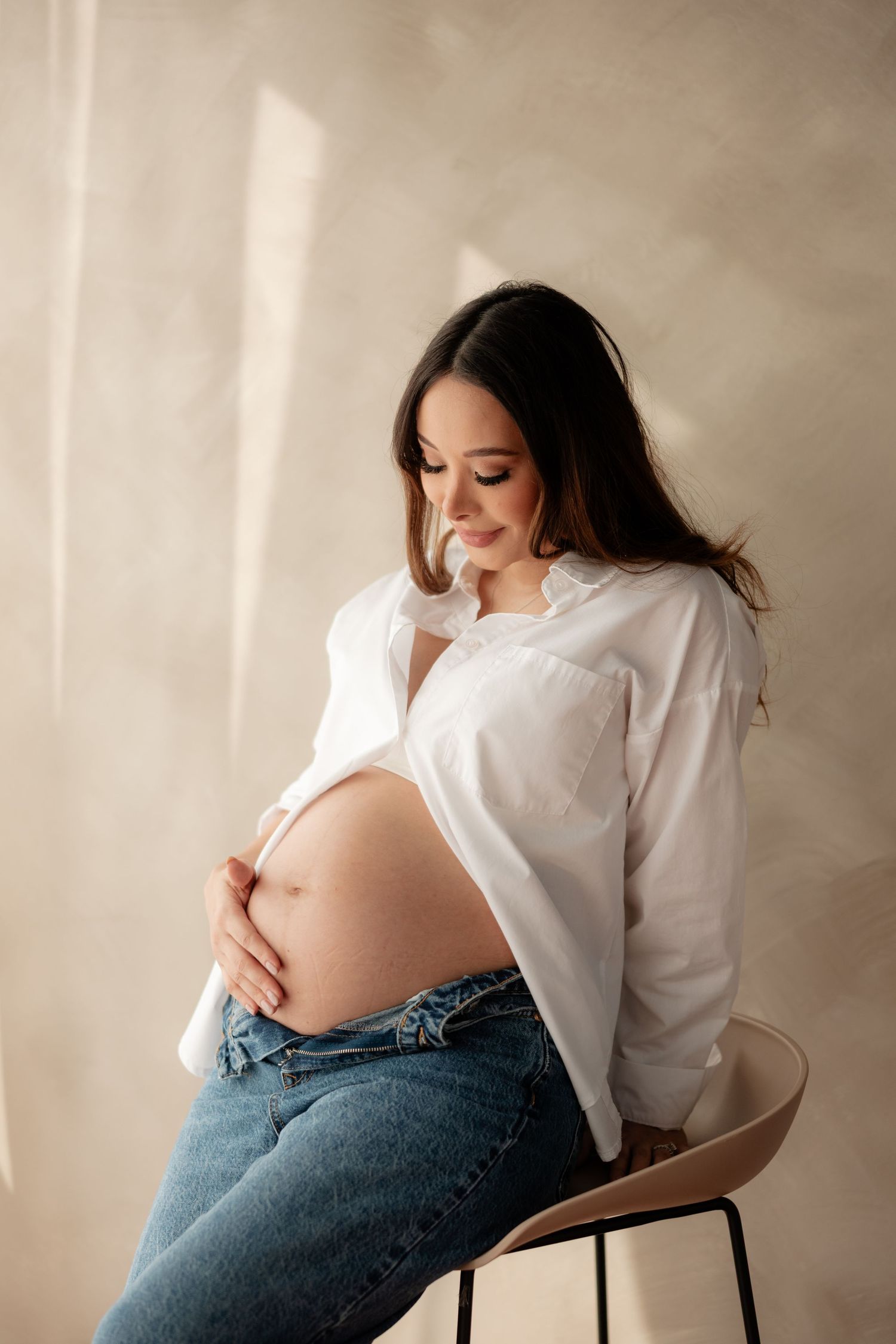 Pregnant woman in white shirt and jeans sitting on stool, tenderly holding her belly.