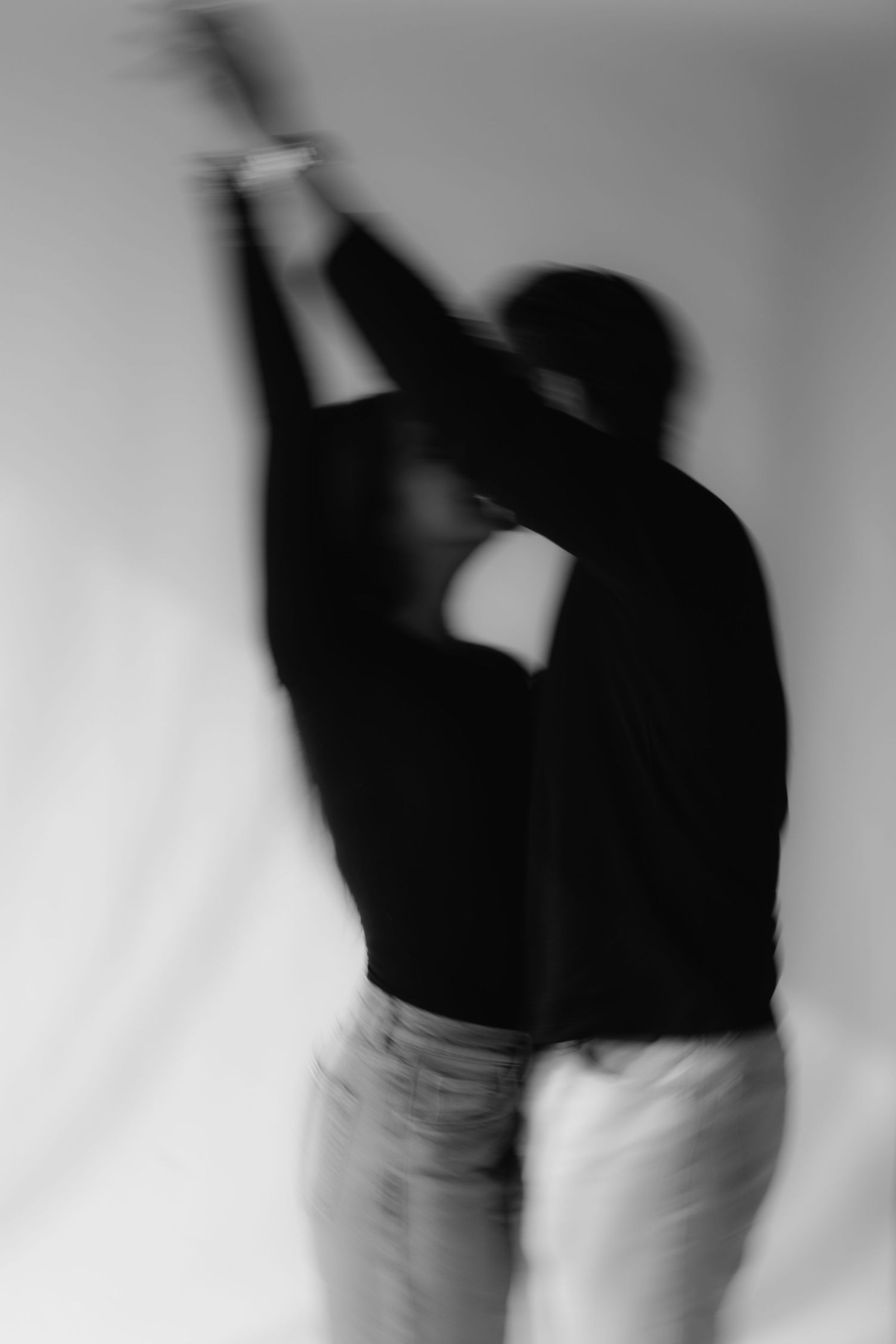 Blurry black and white photo of a person in dark top and light pants reaching arms upward against white backdrop.